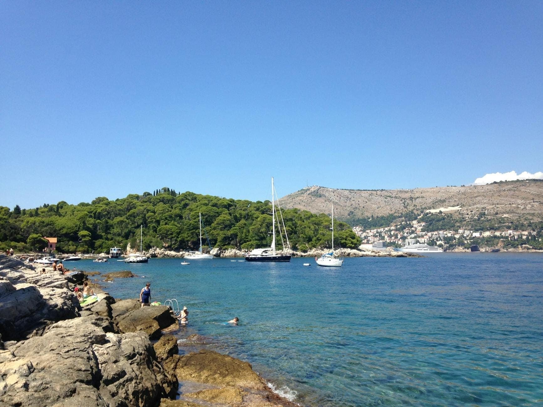 Скалистое побережье с загорающими и купающимися людьми, парусные лодки, стоящие на якоре вблизи острова Локрум, на фоне панорамы Дубровника.