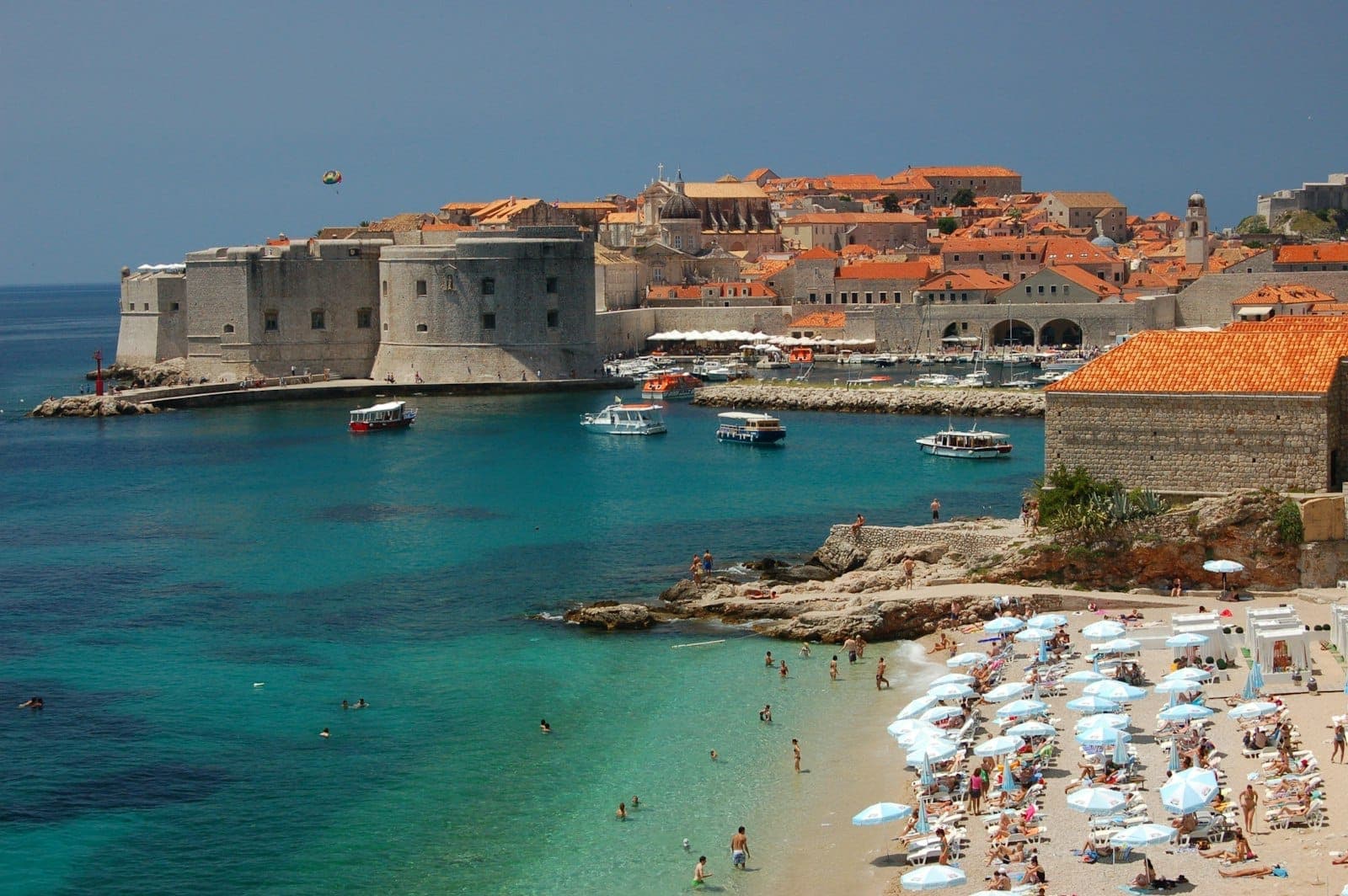 Ein Sandstrand mit Sonnenbadenden und weißen Sonnenschirmen mit Blick auf das türkisblaue Wasser und die alten Stadtmauern von Dubrovnik, Boote nahe am Ufer unter klarem Himmel.