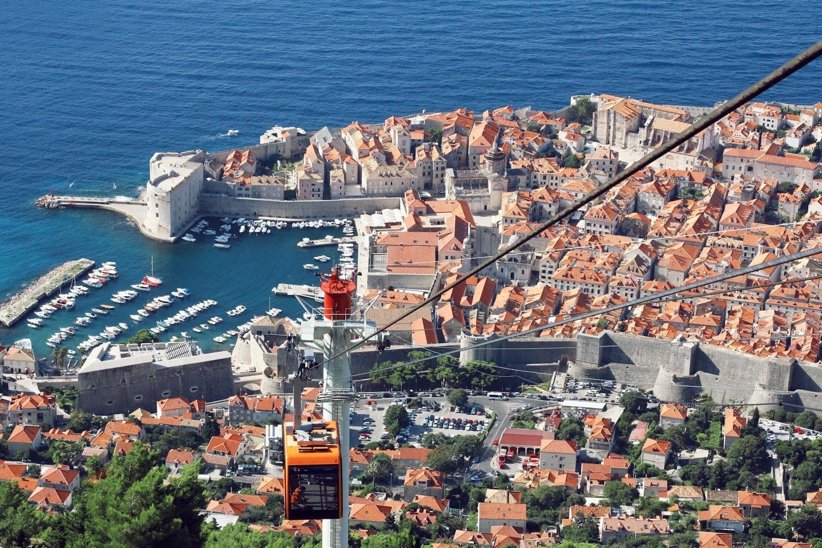 Vista aérea da cidade antiga e do porto de Dubrovnik, com uma cabine laranja do teleférico em primeiro plano subindo o monte Srđ.
