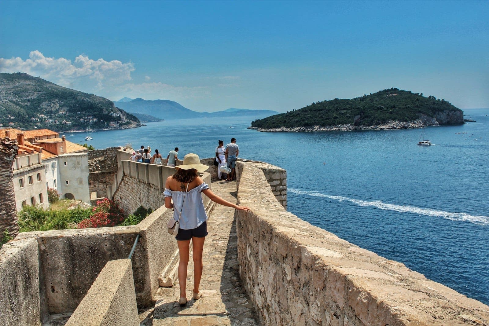 Touristen spazieren auf den Stadtmauern von Dubrovnik mit Blick auf die Adria und eine nahegelegene Insel bei strahlendem Sonnenschein.