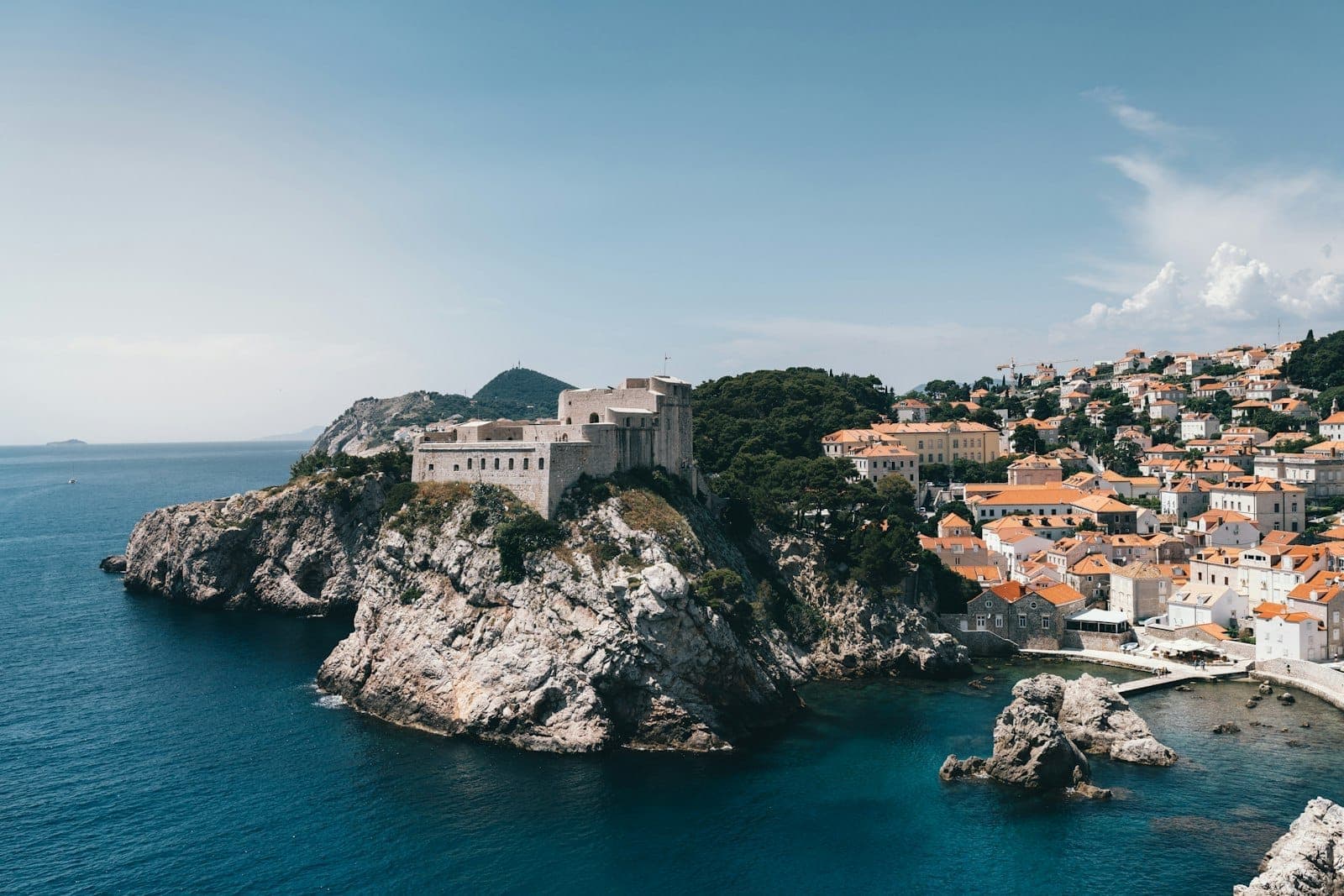 View of Dubrovnik's stone city walls and old town on the Adriatic coast, with clear skies, cliffs and terracotta rooftops.