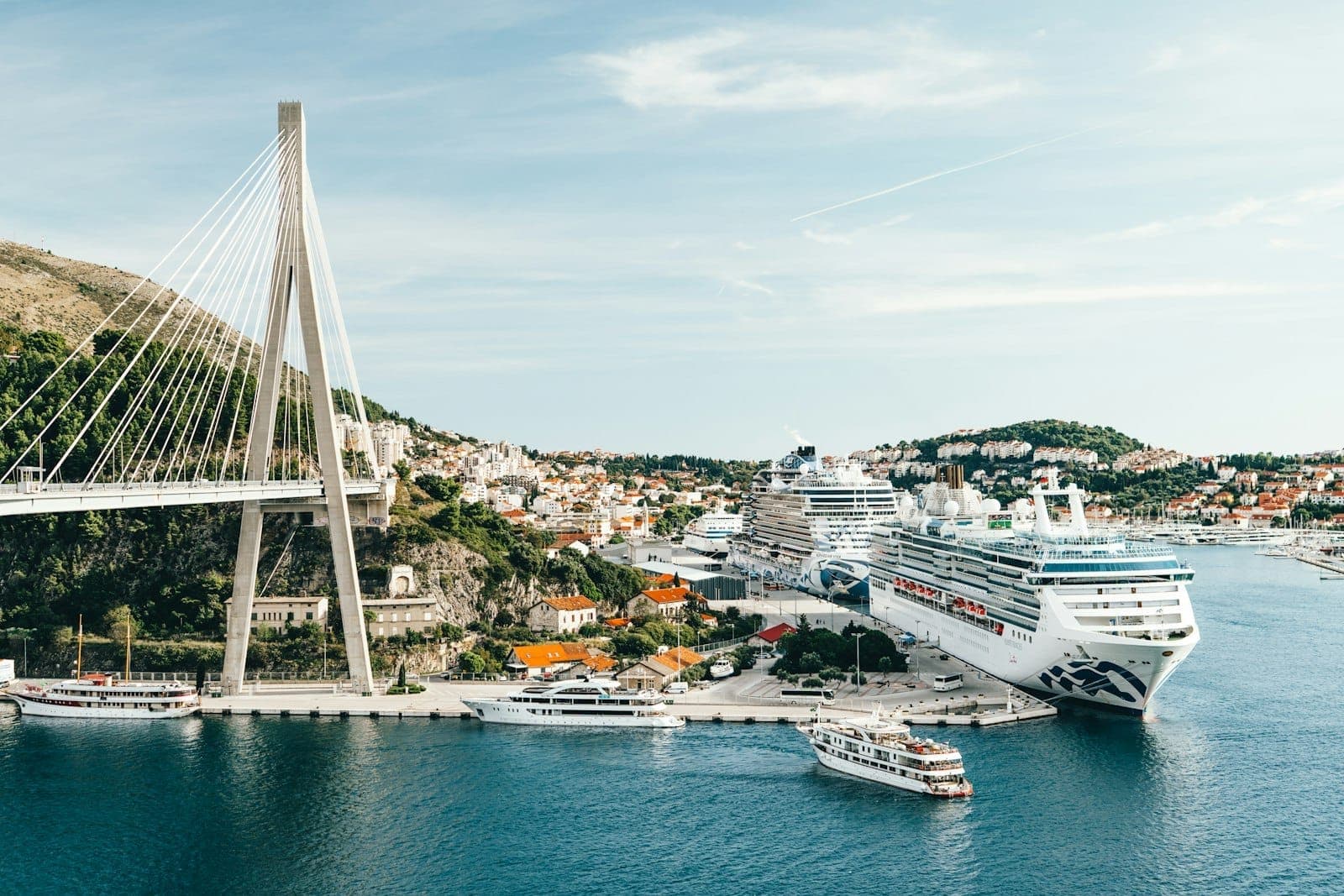 Large cruise ships docked at Dubrovnik’s main port near a cable-stayed bridge, with city buildings and hills in the background.