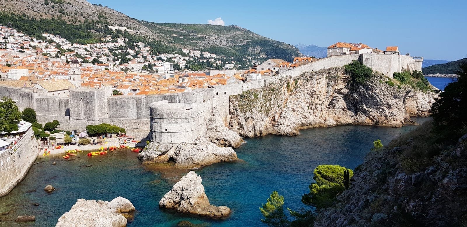 Panoramablick auf Dubrovniks Altstadt mit Stadtmauern, Felsküste, türkisblauem Wasser und orangefarbenen Dächern vor grünen Hügeln unter blauem Himmel.