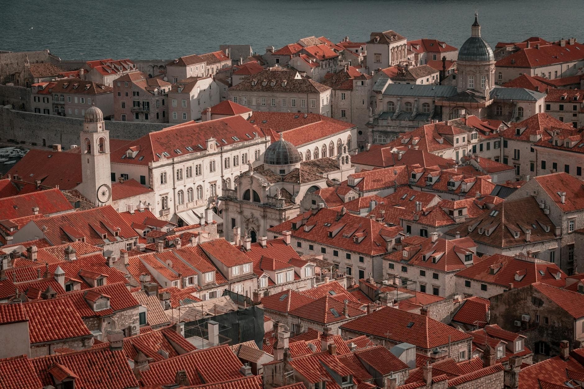 Pemandangan atap-atap Kota Tua Dubrovnik dengan bangunan bersejarah, kubah, dan menara gereja yang padat di dalam tembok kota.