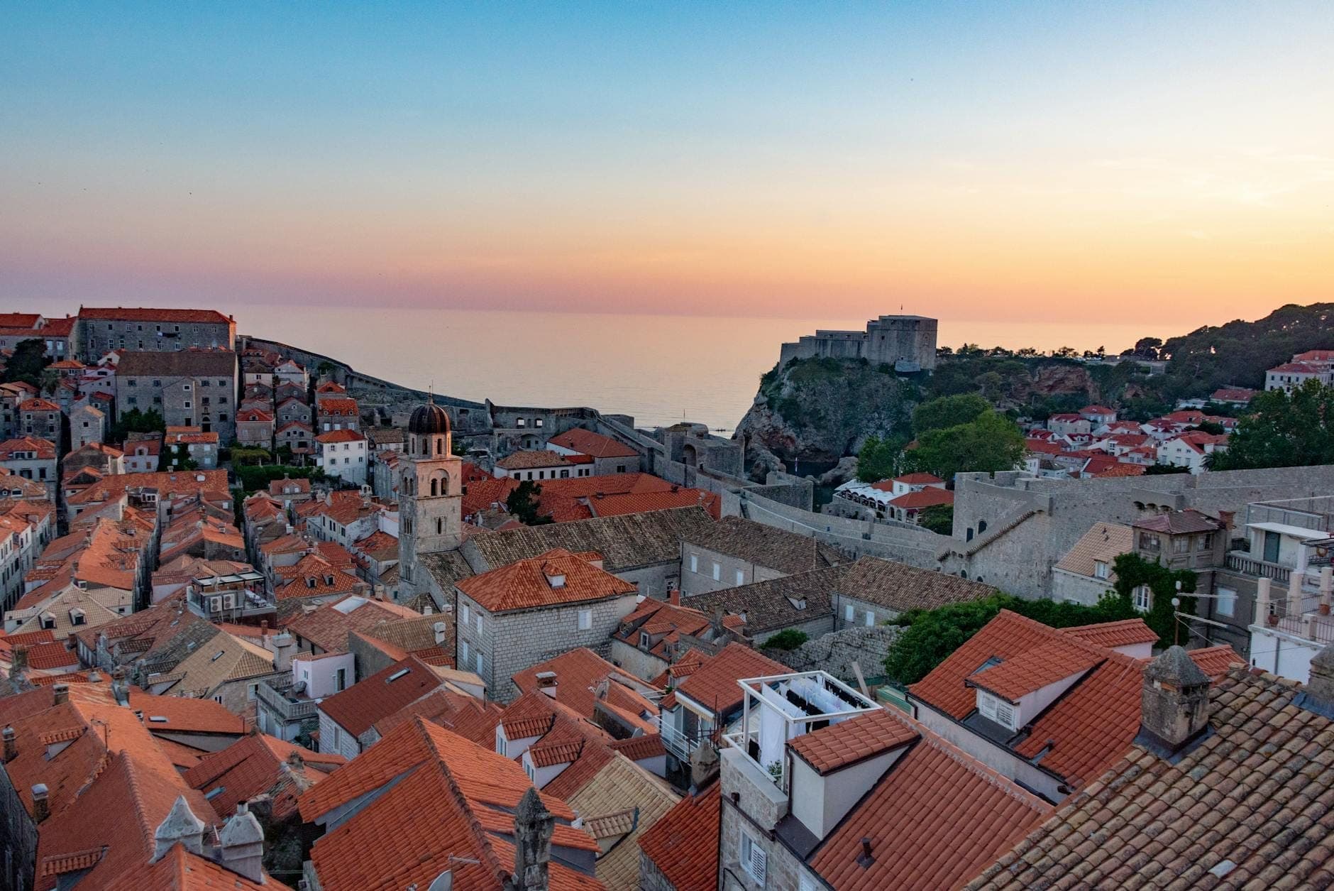 Pemandangan matahari terbenam yang romantis di atas tembok dan atap Kota Tua Dubrovnik dengan laut di latar belakang.