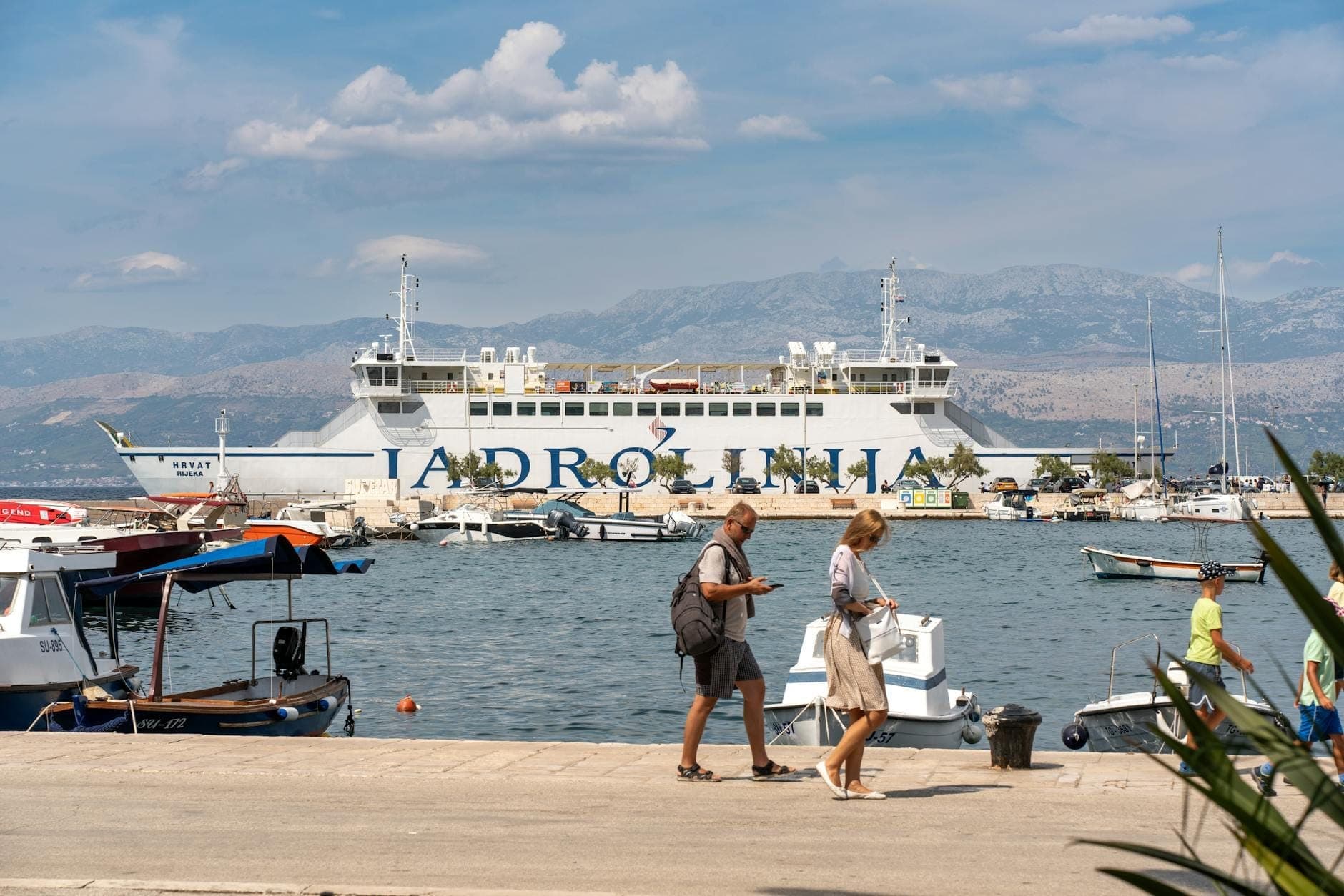 Große Jadrolinija-Fähre liegt in einem Hafen, Menschen spazieren entlang des Piers und Boote sind im Hintergrund zu sehen.