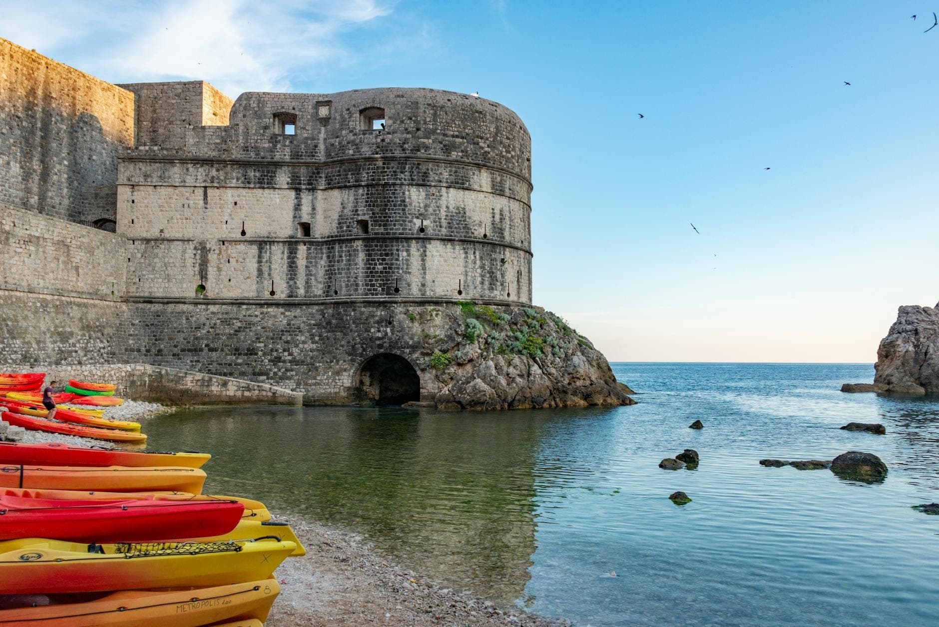 Bunte Kajaks am Ufer neben Dubrovniks alten Steinmauern, dahinter die Adria und ein strahlend blauer Himmel.