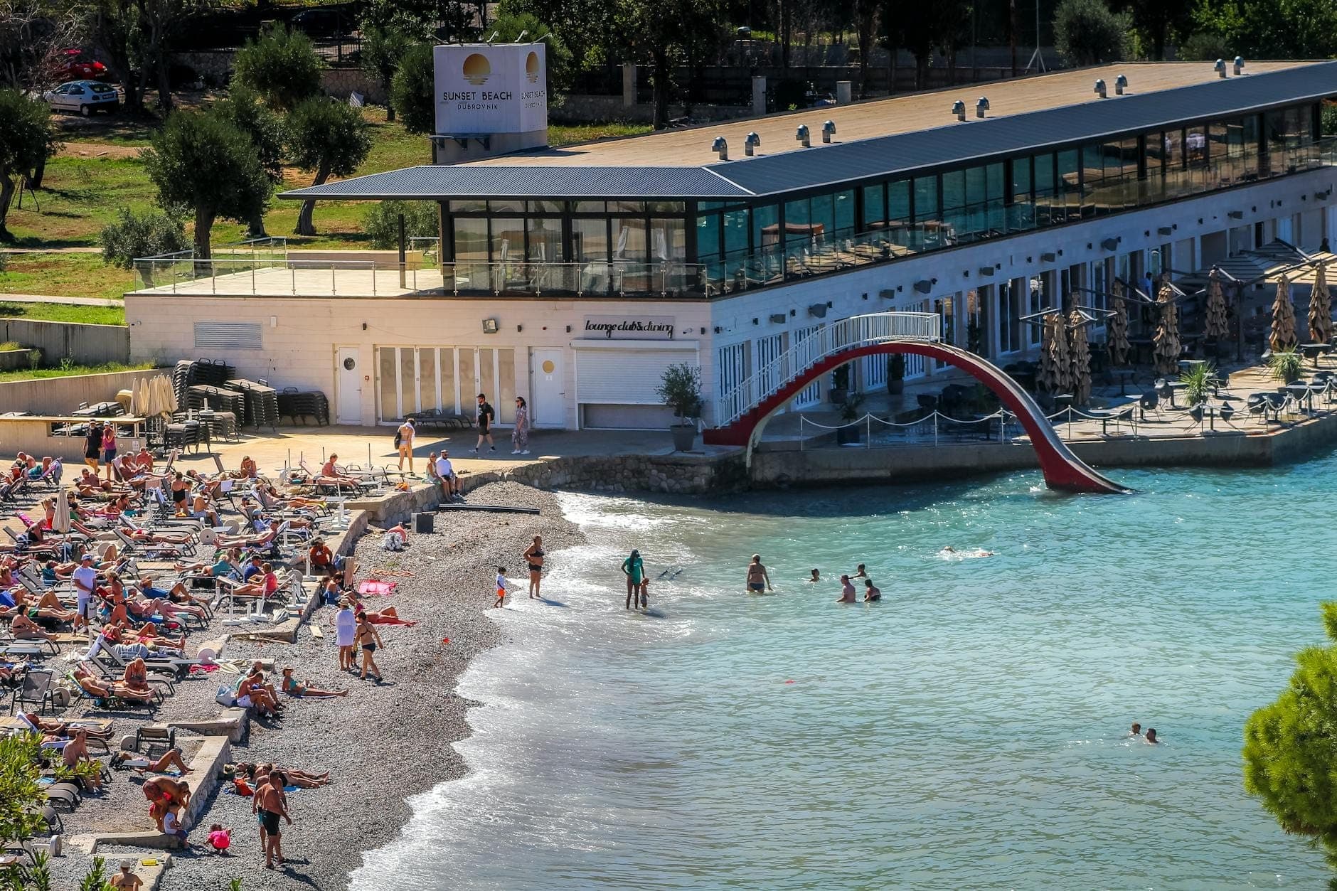 บีชคลับใน Dubrovnik ที่คึกคักพร้อมเตียงผึ่งแดด ผู้คนพักผ่อน ว่ายน้ำ และอาคารบาร์ทันสมัยที่มองเห็นทะเลใสสวยงาม