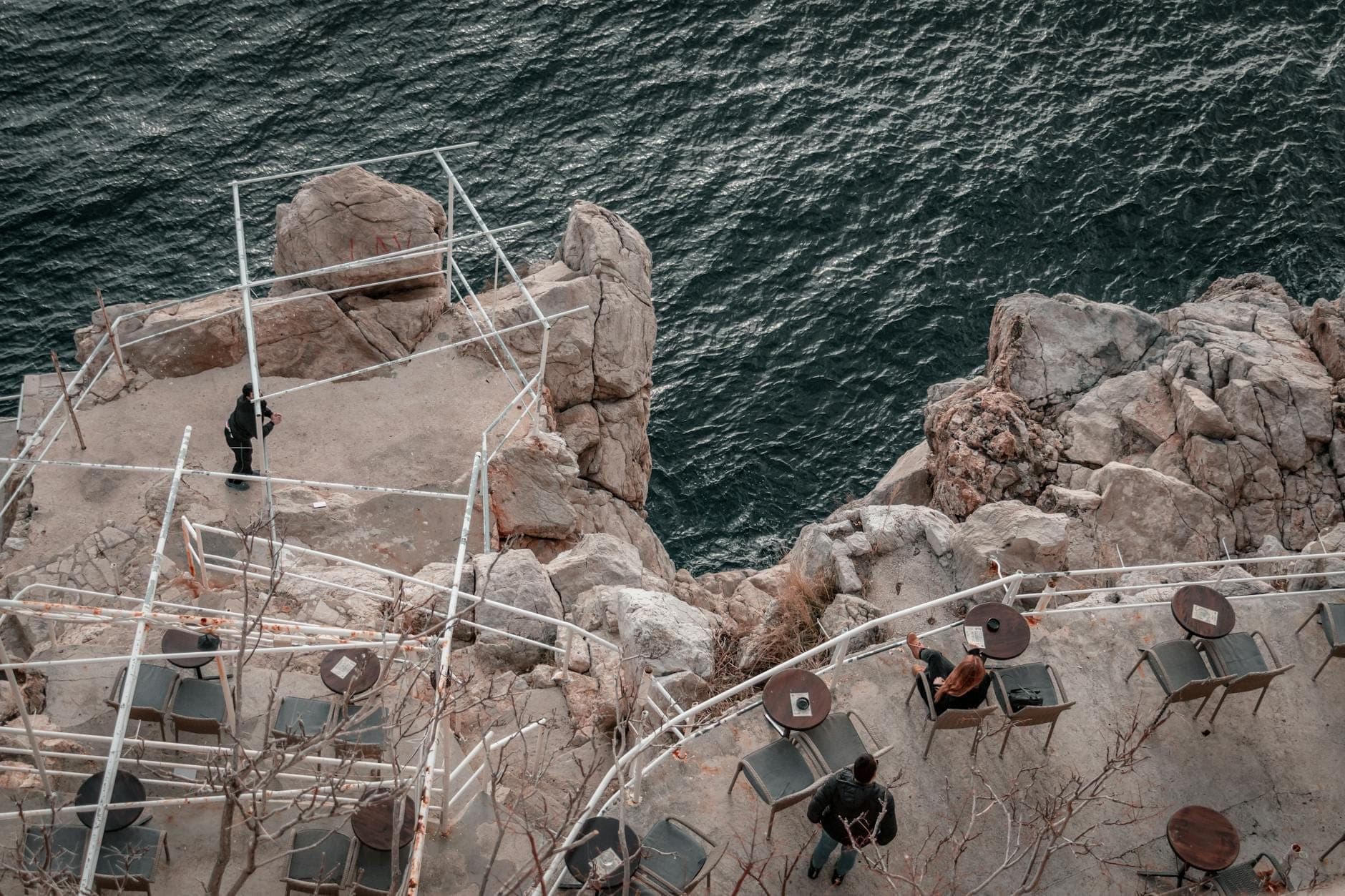 โต๊ะและเก้าอี้บนระเบียงบาร์ริมหน้าผาที่มองเห็นมหาสมุทร พร้อมกับผู้คนที่เพลิดเพลินกับวิวอันตระการตาบนขอบหินใน Dubrovnik