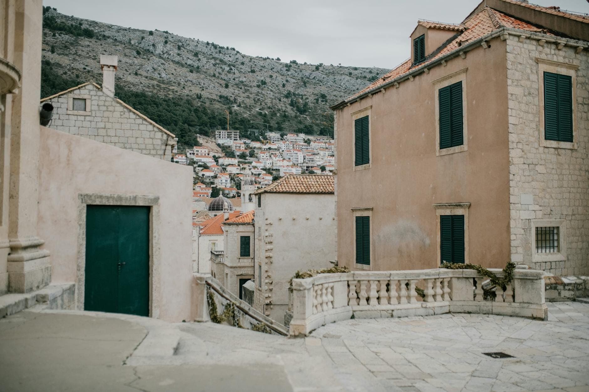 Spokojna ulica mieszkalna w Dubrowniku z kamiennymi domami, zielonymi okiennicami i domami na zboczu wzgórza w tle, przywołująca atmosferę lokalnej dzielnicy.