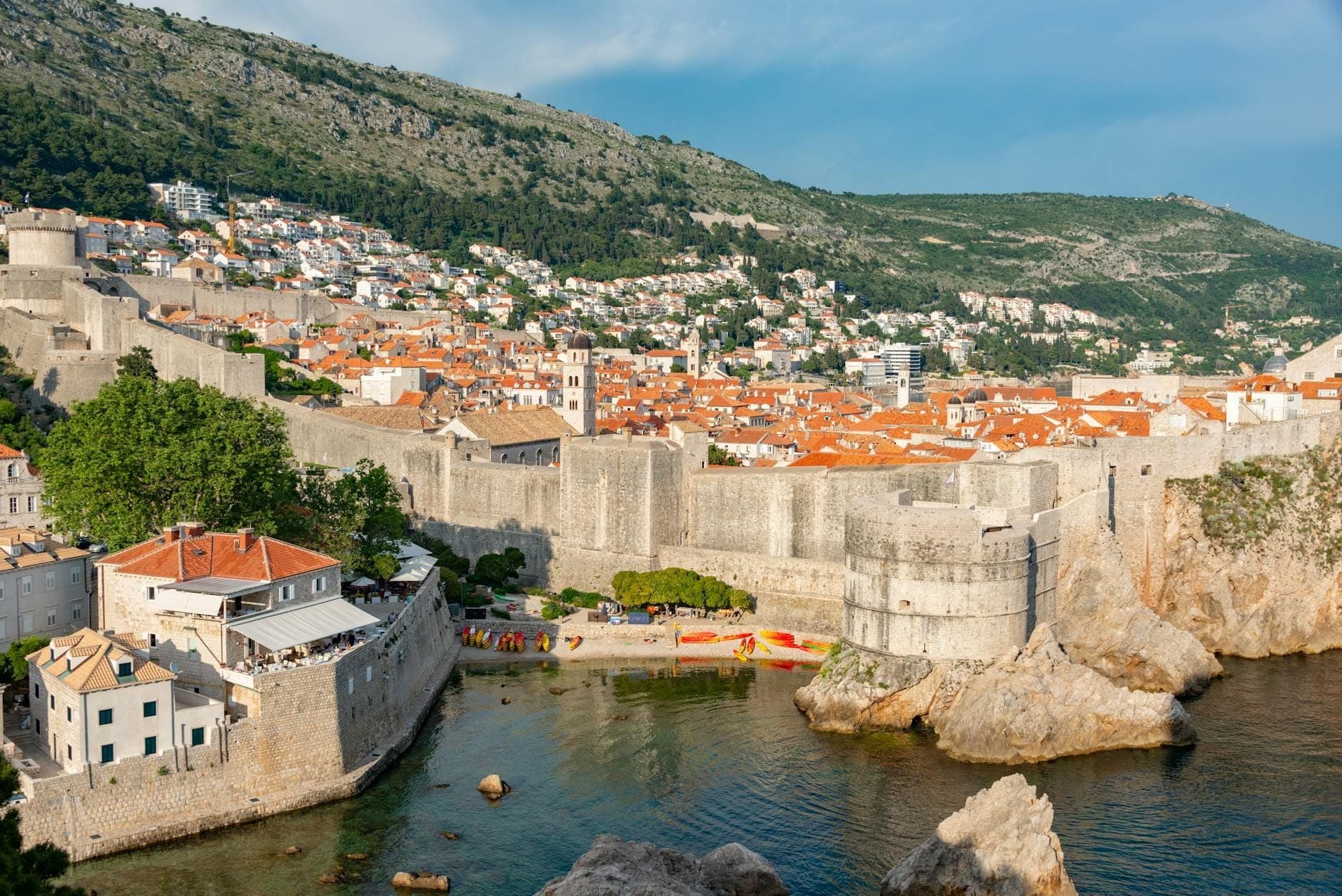 Szeroki widok na mury miejskie Dubrovnika z basztami i budynkami starego miasta z czerwonymi dachami wtulonymi w zbocze wzgórza, z widokiem na Morze Adriatyckie.