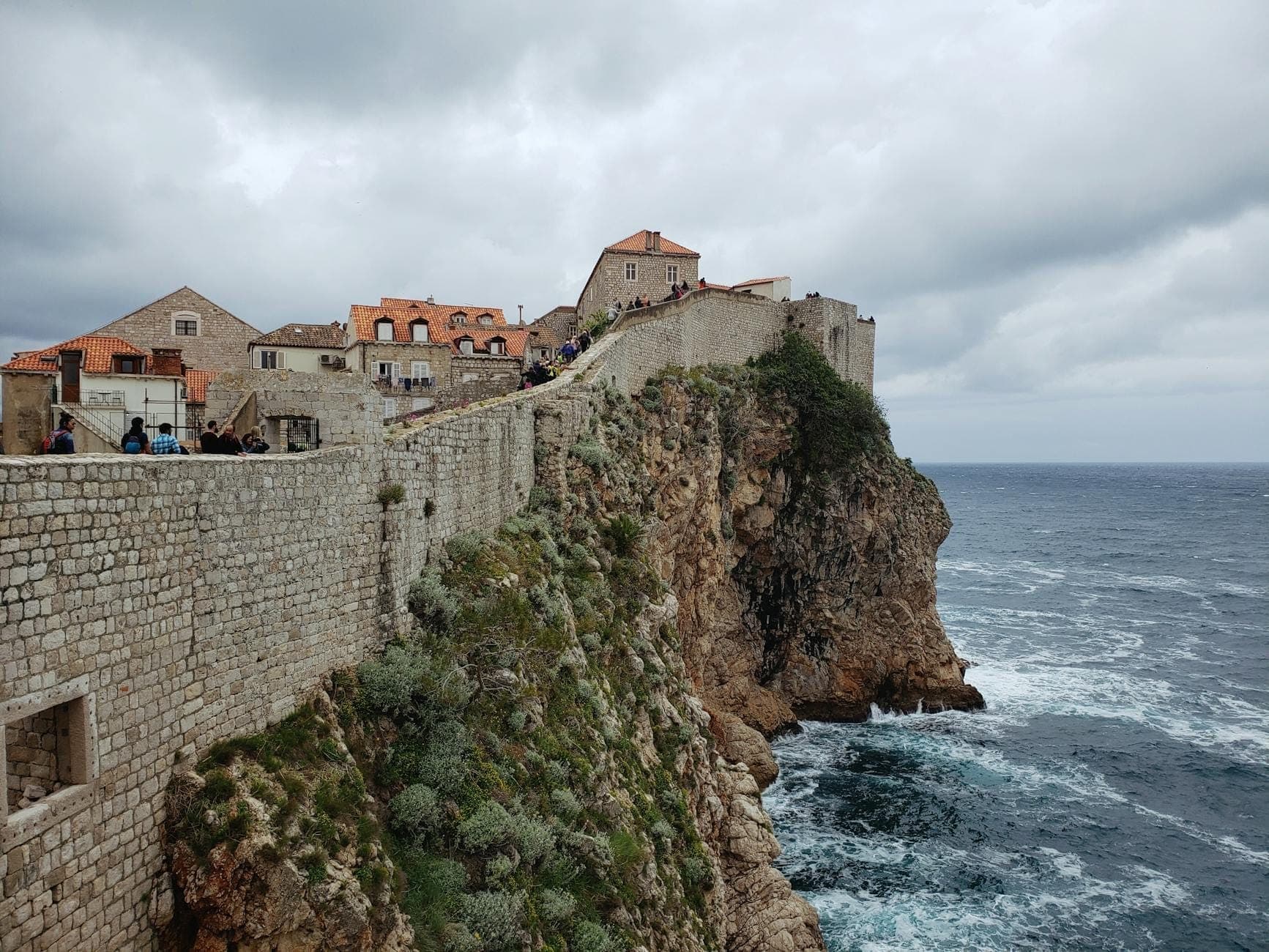 Vista de las murallas históricas de la Ciudad de Dubrovnik a lo largo de un dramático acantilado sobre el mar Adriático, con edificios de tejados naranjas visibles.