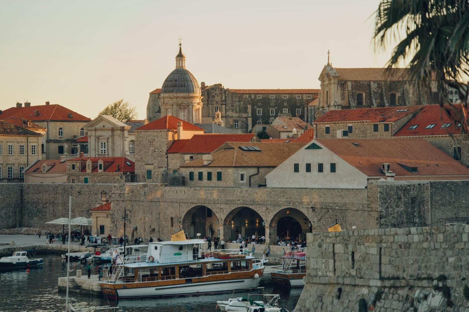ท่าเรือดูบรอฟนิกพร้อมเรือ กำแพงหินของเมือง และอาคารประวัติศาสตร์ยามพระอาทิตย์ตกดิน สะท้อนบรรยากาศการเดินทางและวิถีชีวิตท้องถิ่น