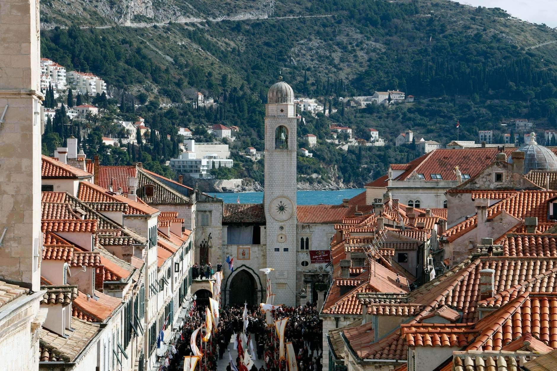 Blick über Dubrovniks ikonische Altstadt mit orangefarbenen Dächern, einem markanten Glockenturm und einer feiernden Menschenmenge auf der Hauptstraße.