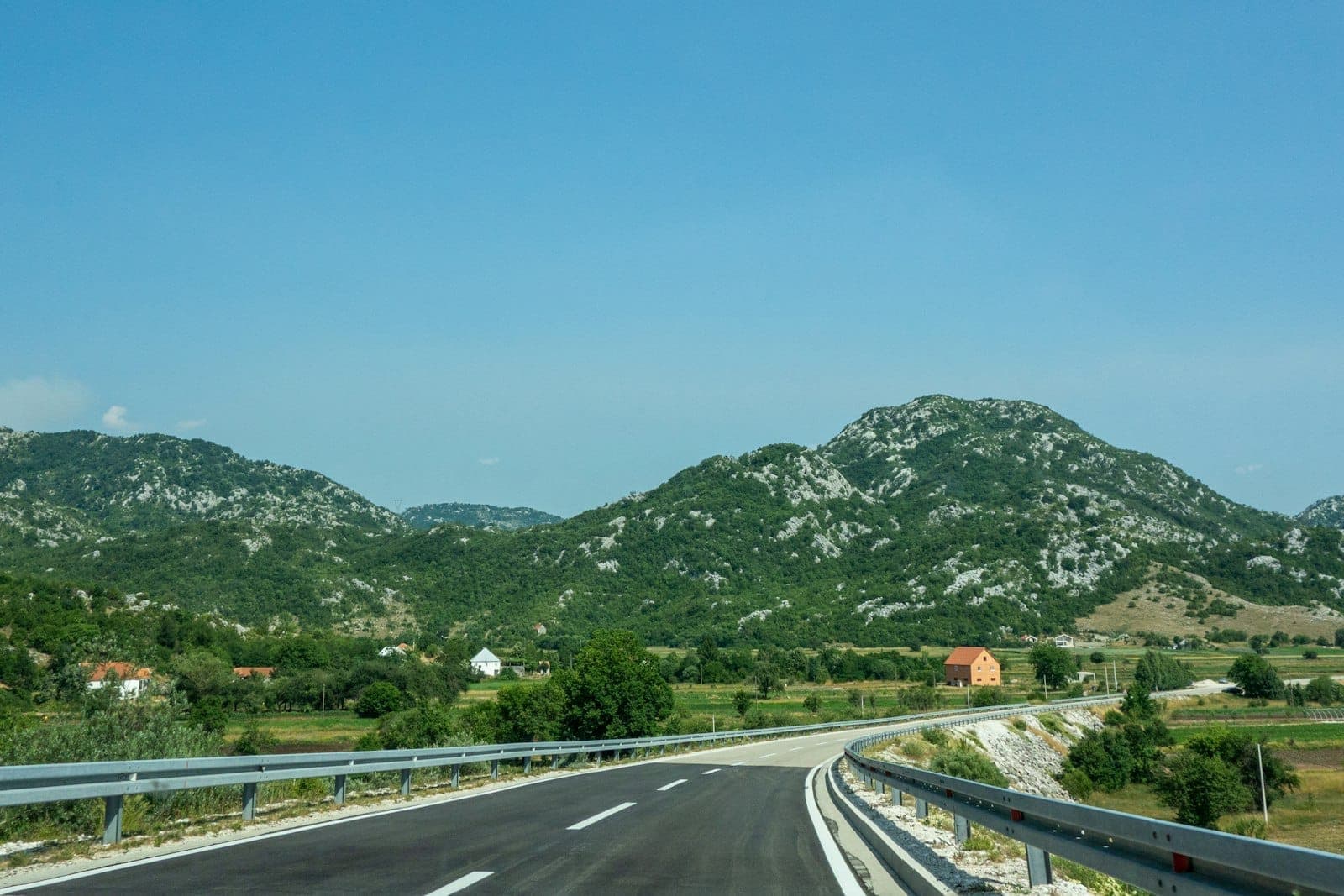 Une route goudronnée lisse avec des glissières de sécurité qui serpente vers des collines et des montagnes verdoyantes, évoquant le trajet vers un poste frontière.