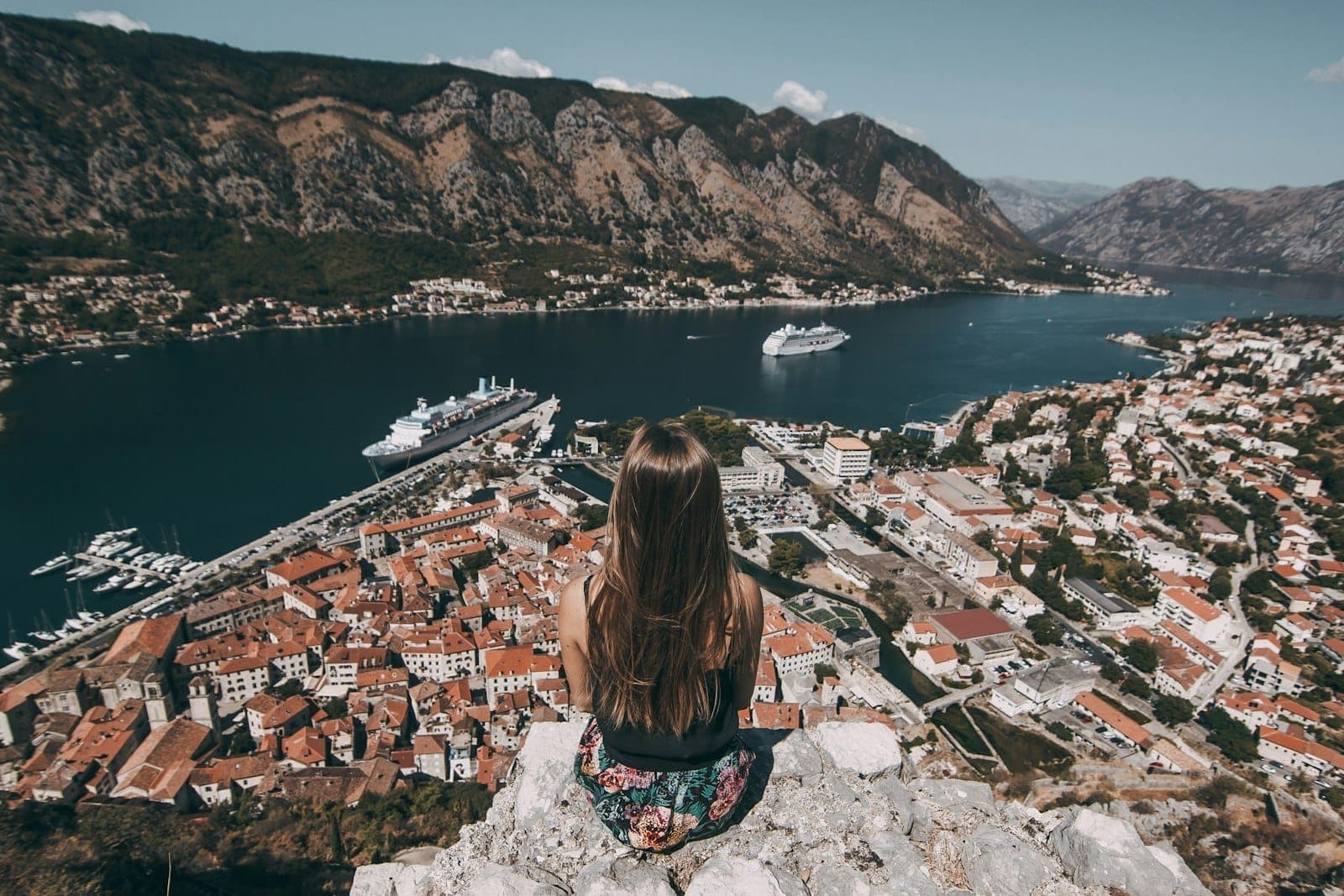 Une femme assise sur des rochers surplombant la Baie de Kotor, avec des toits en terre cuite, des paquebots de croisière et des montagnes spectaculaires sous un ciel lumineux de midi.