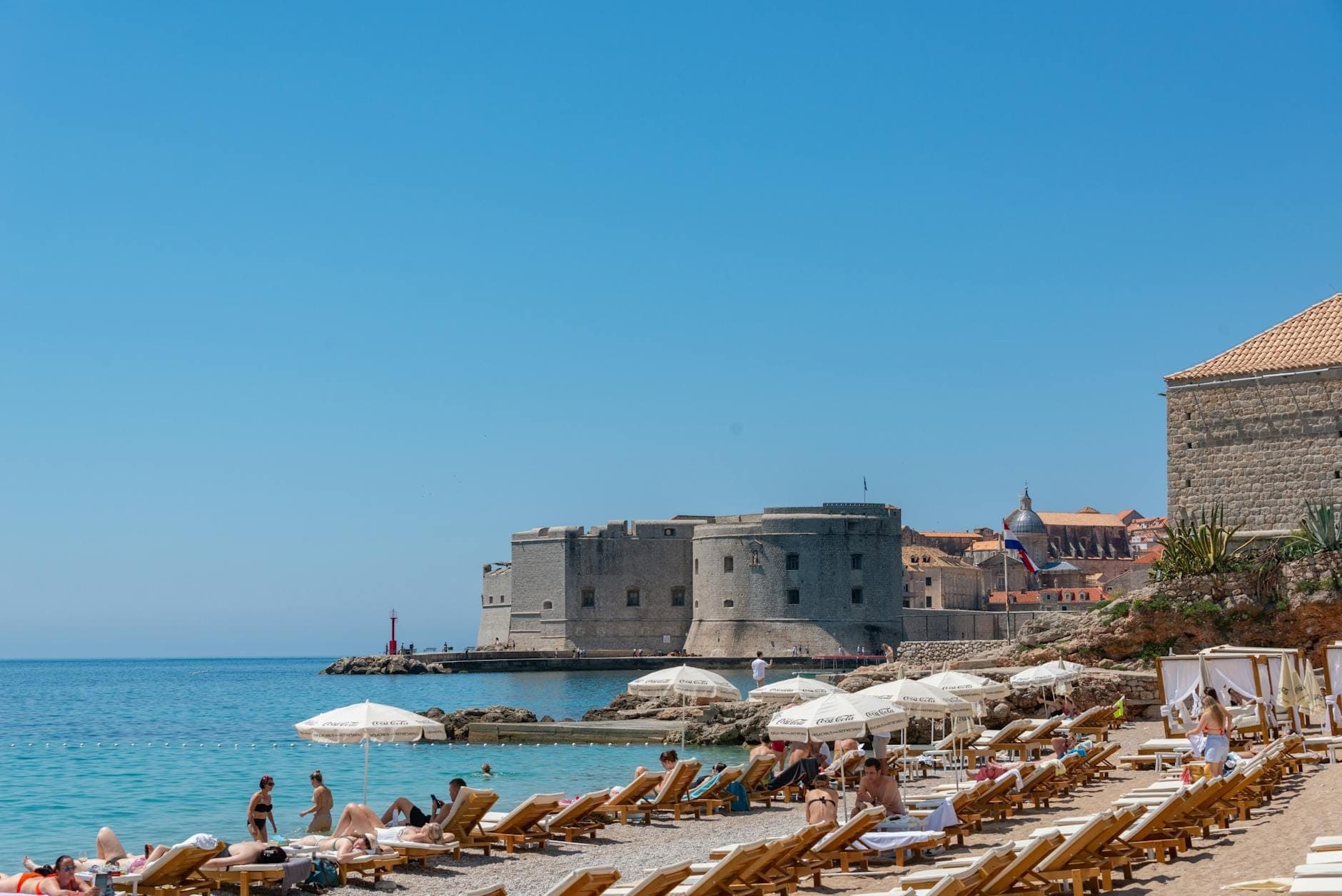 Blick auf die Altstadtmauern von Dubrovnik mit Sonnenbadenden an einem wenig belebten Sandstrand mit Sonnenschirmen und klarem blauem Meer unter strahlend blauem Himmel.