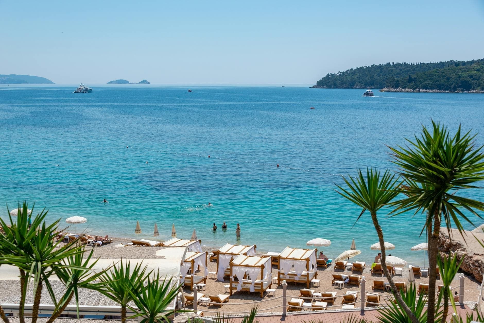 Sonniger Strand von Dubrovnik mit Strandhütten und schwimmenden Menschen im klaren blauen Adriatischen Meer, mit Booten in der Ferne und einer üppig grünen Insel im Hintergrund.