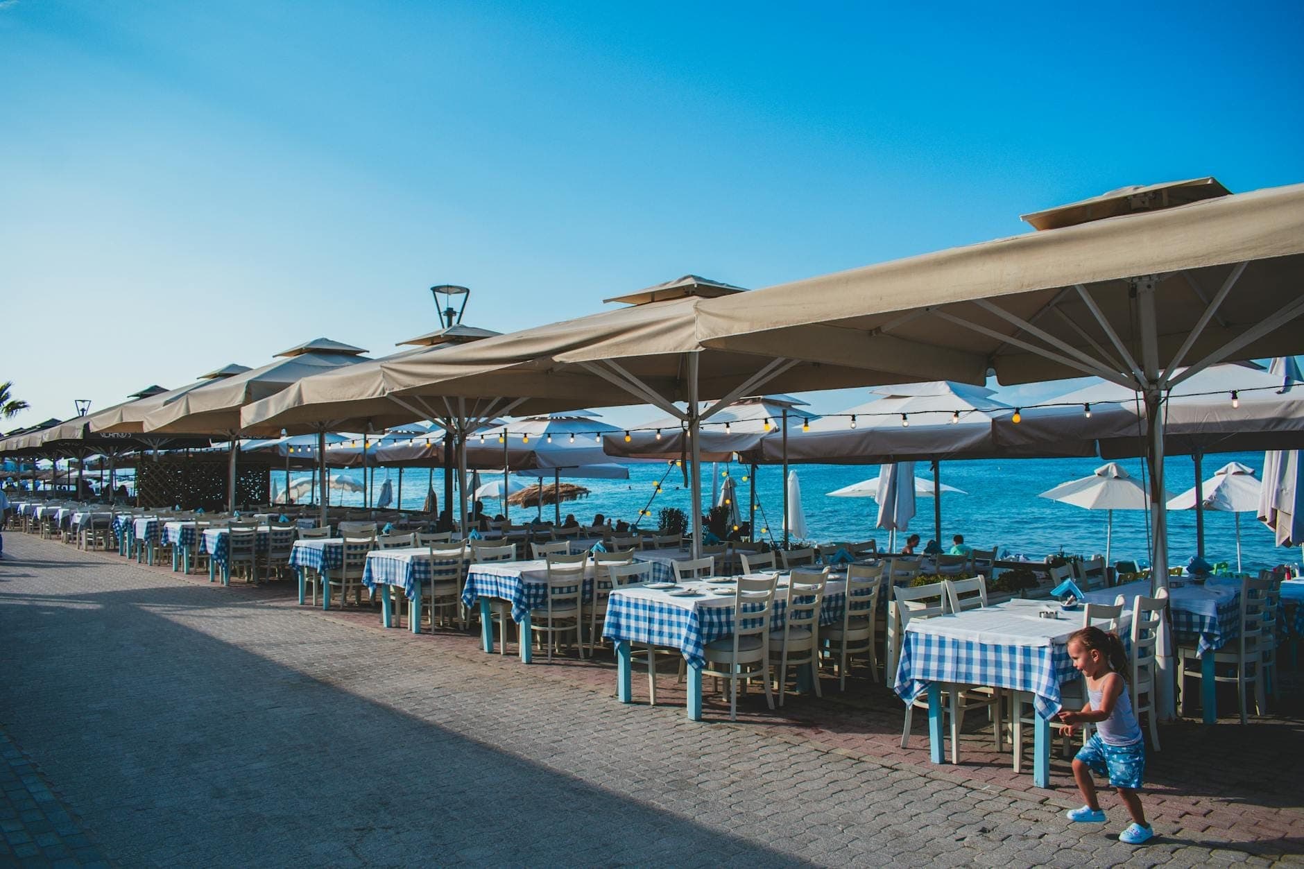 Restaurante ao ar livre à beira-mar em Dubrovnik com toalhas de mesa azuis e brancas, guarda-sóis e uma criança perto das mesas sob um céu azul limpo.