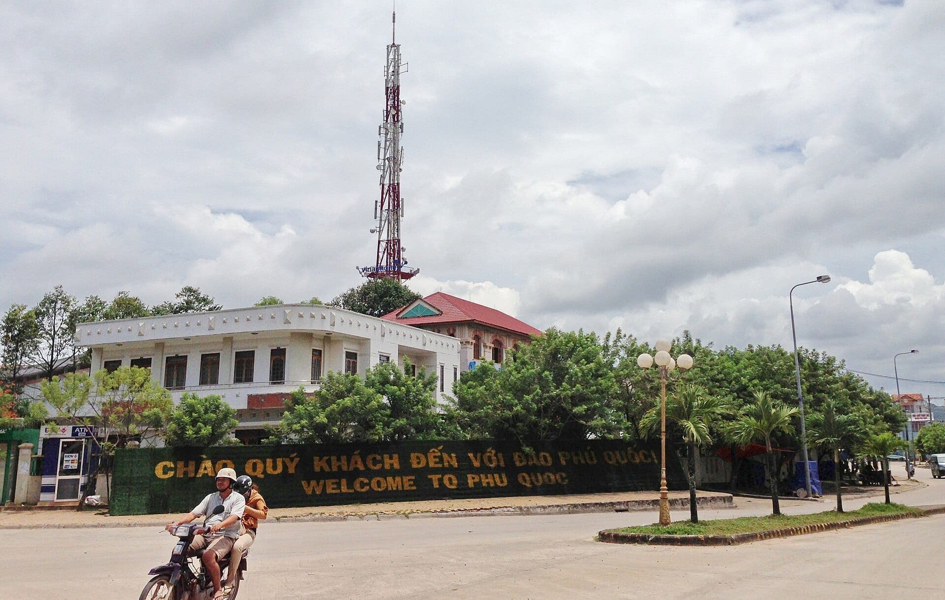 Panneau de bienvenue à Duong Dong, Phu Quoc, avec des motocyclistes passant devant des bâtiments locaux en arrière-plan