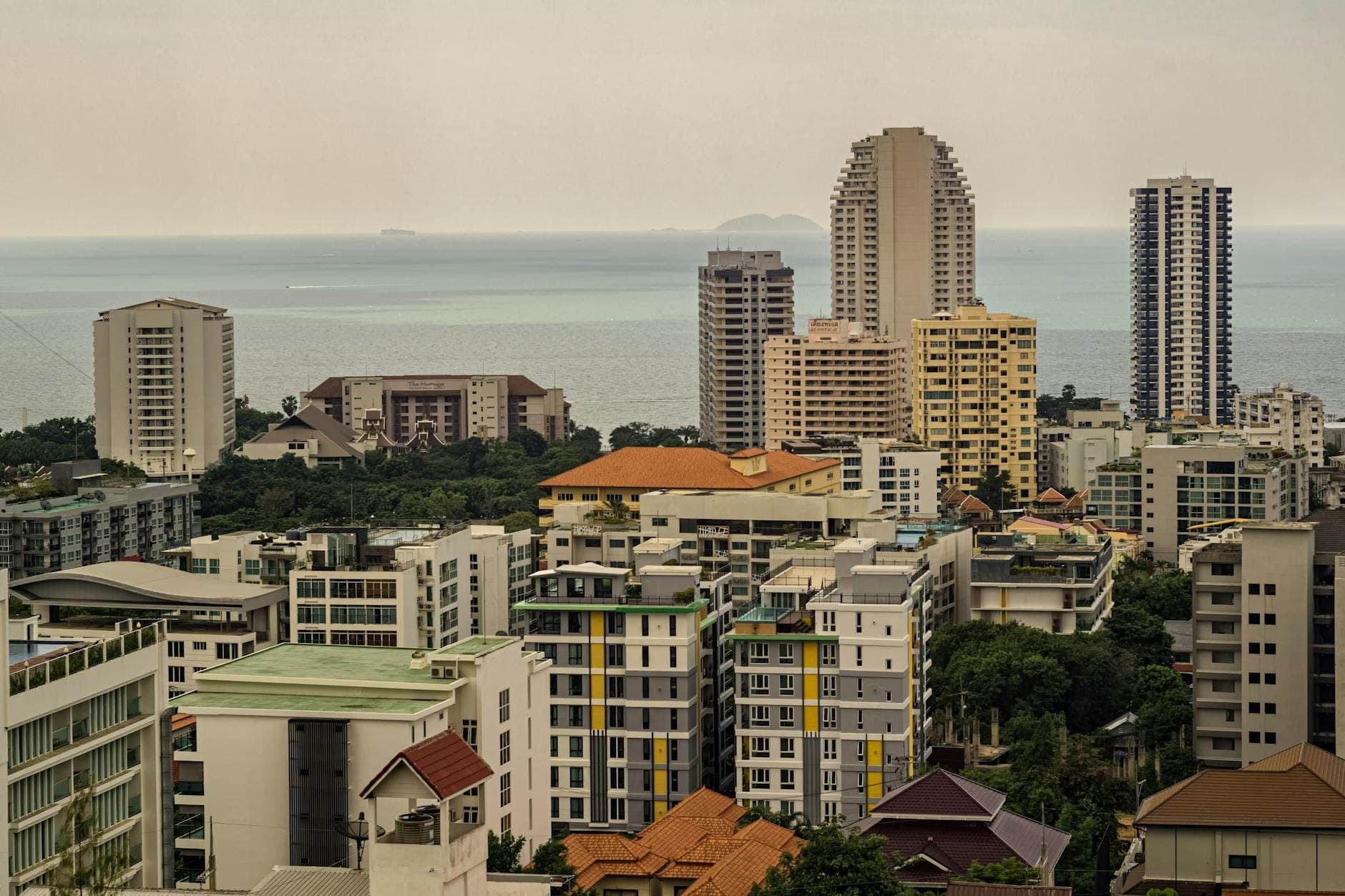 A landscape view of low-rise buildings, houses, and greenery in East Pattaya with the sea and distant islands visible under a hazy sky.