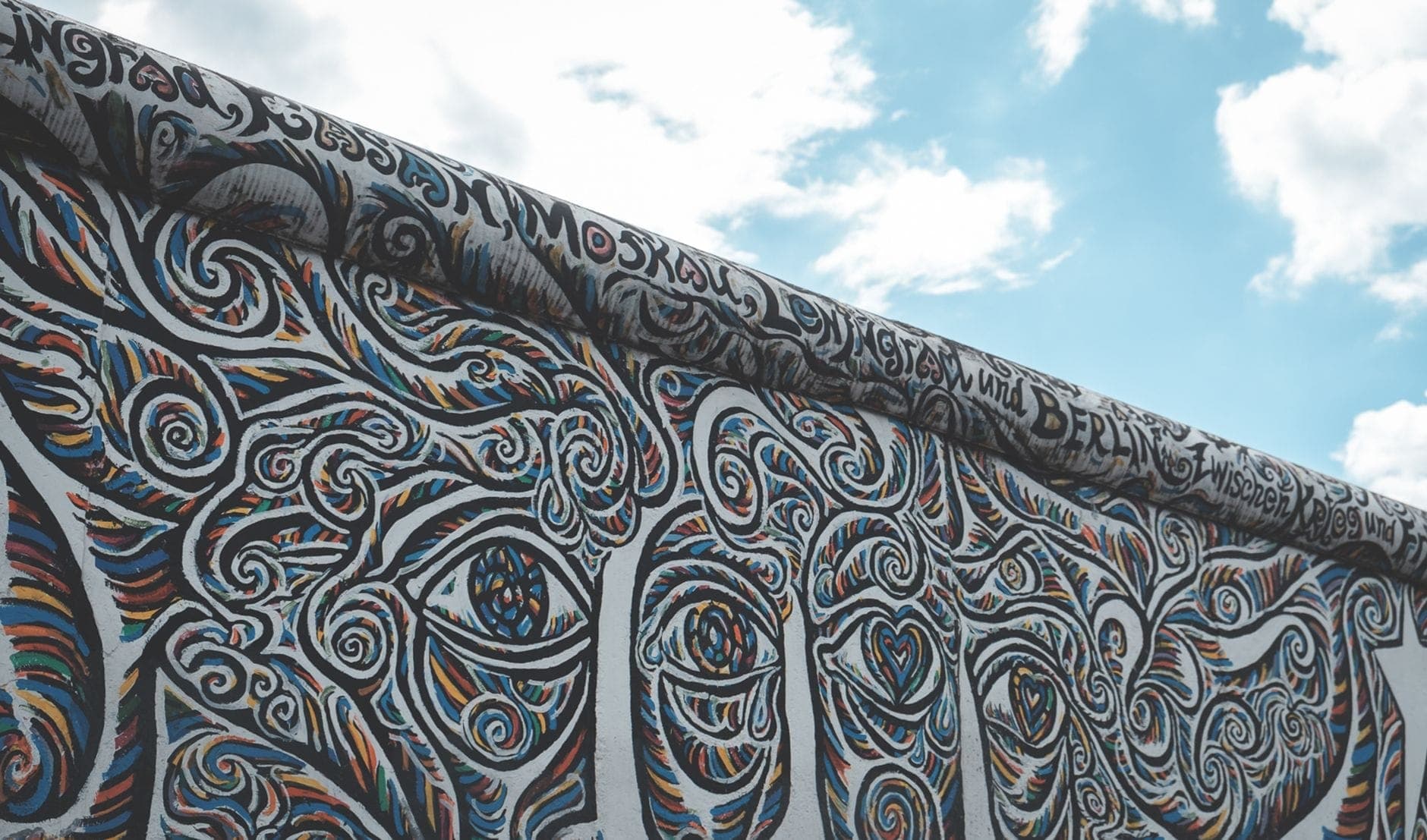 กำแพงเบอร์ลินส่วน East Side Gallery เพ้นท์ลวดลายสีสันสดใสใต้ท้องฟ้าสีฟ้าและเมฆ