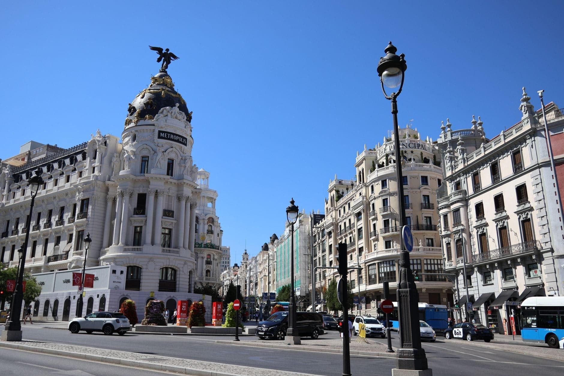 Широкоугольный вид Edificio Metrópolis на углу Calle de Alcalá и Гран-Виа в Мадриде на фоне ясного голубого неба — виден нарядный купол и оживлённое городское окружение.