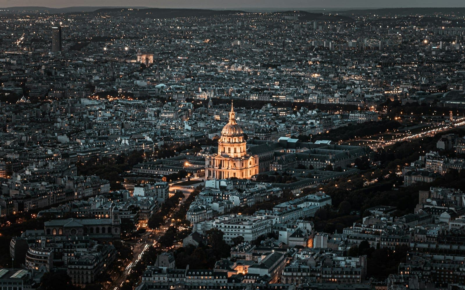 Aerial view of Paris at dusk with the illuminated golden dome of Les Invalides standing out in the center and cityscape stretching into the distance.