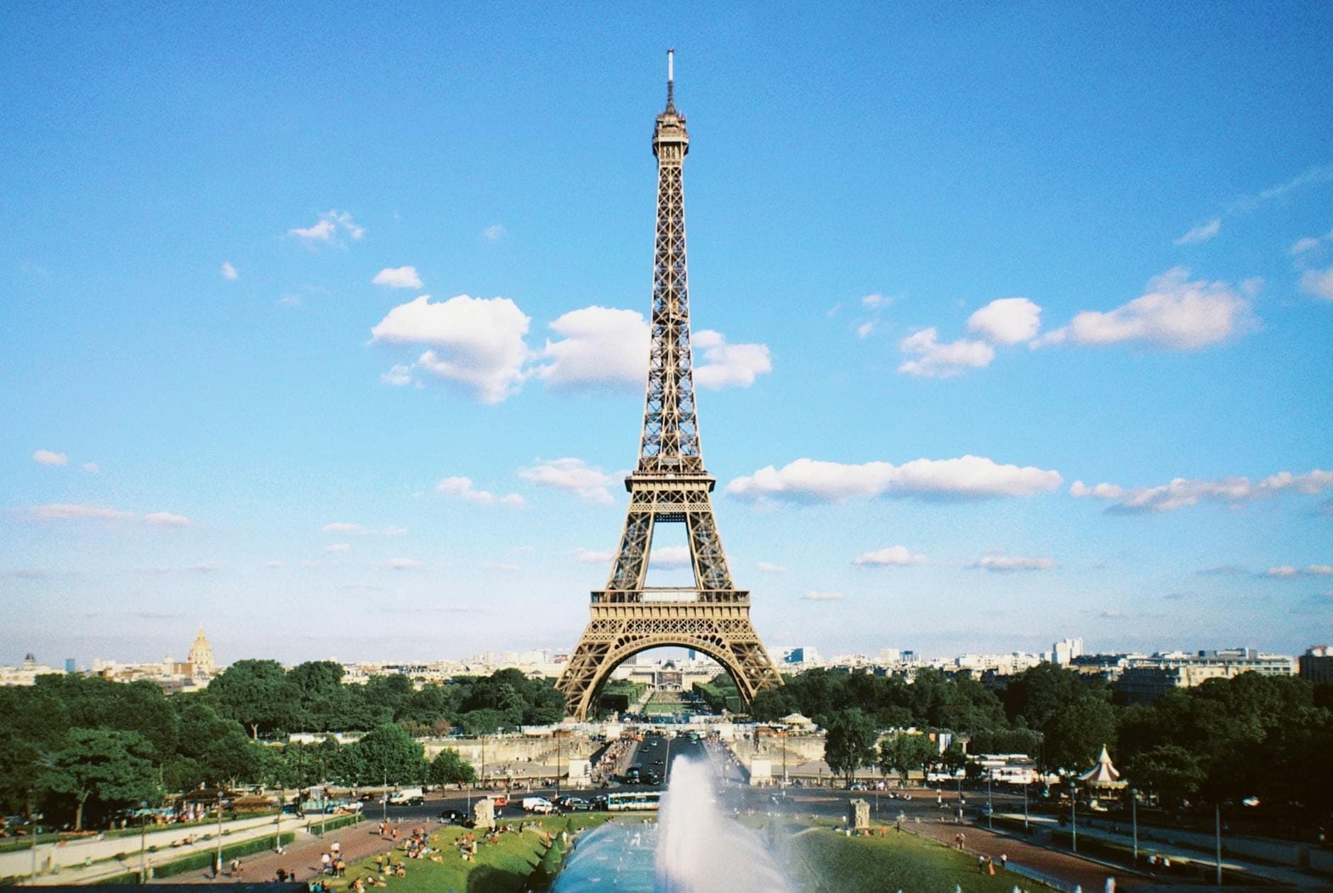 Toàn cảnh Tháp Eiffel trên nền trời xanh với vườn cây xanh mướt, đài phun nước và du khách thư thái tận hưởng không gian Paris.
