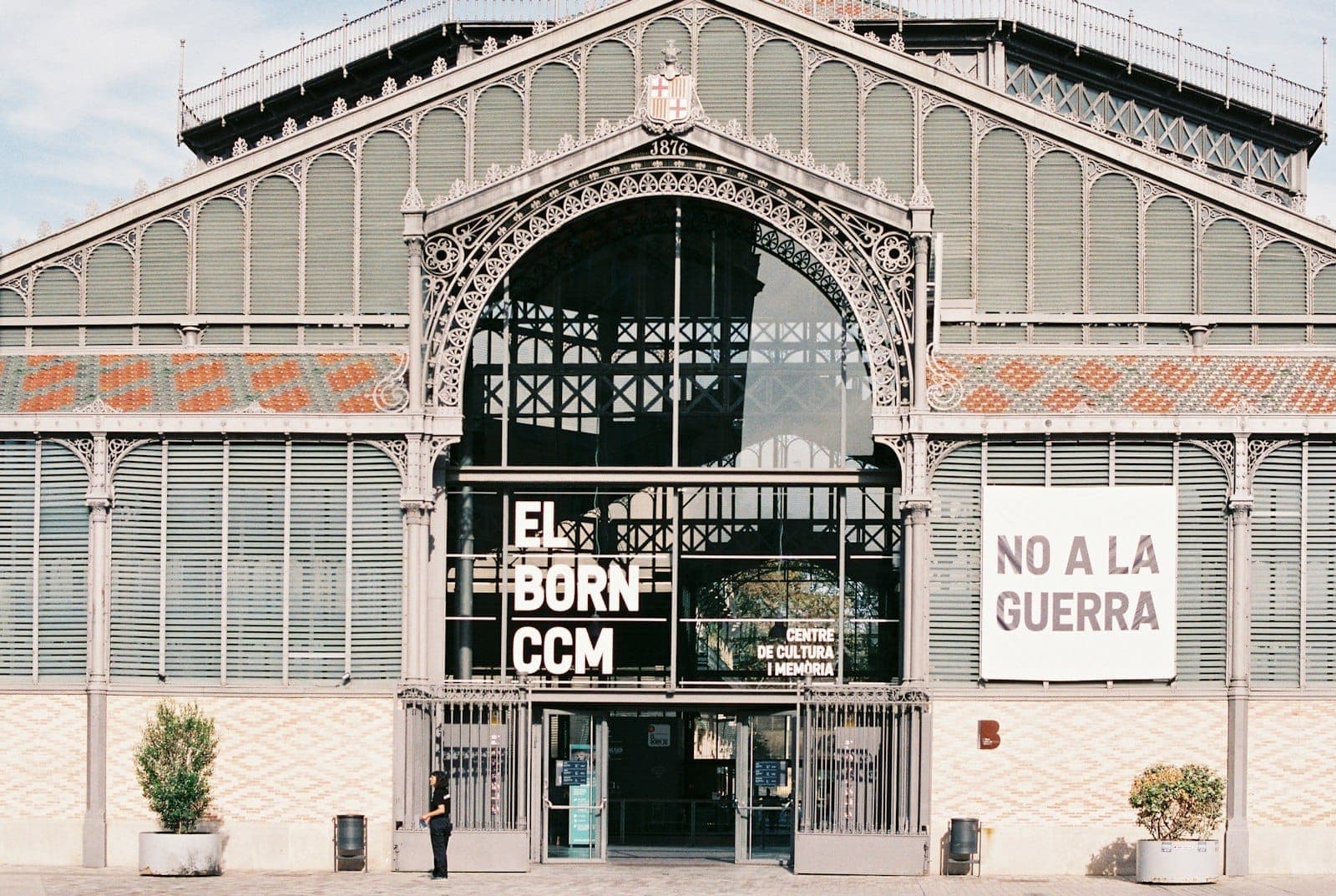 มุมมองด้านหน้าของ El Born Centre de Cultura i Memòria พร้อมสถาปัตยกรรมเหล็กประดับประดา ป้ายแบนเนอร์ และผู้คนหนึ่งคนบริเวณทางเข้า