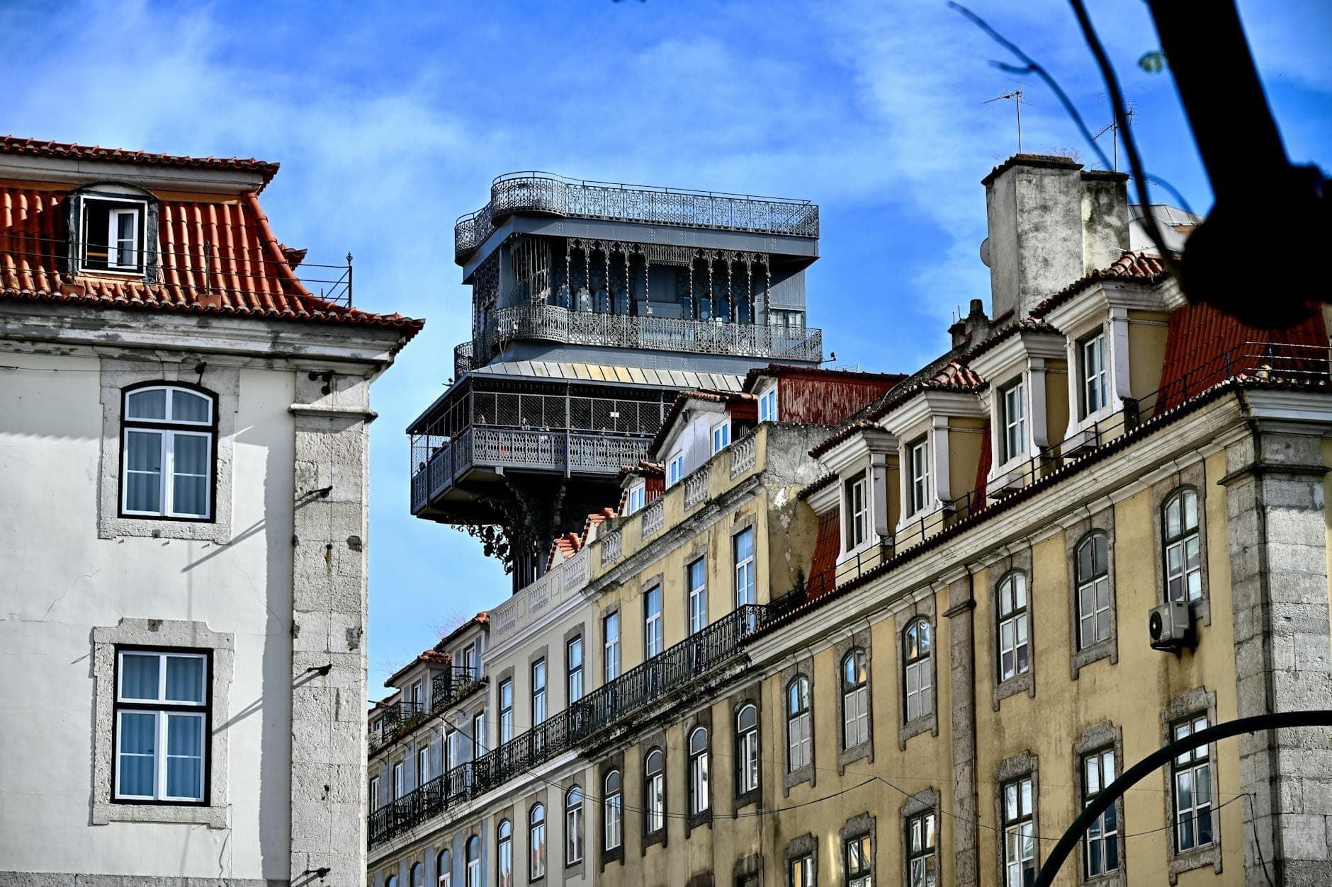 A torre de ferro do Elevador de Santa Justa se ergue acima dos prédios históricos de Lisboa sob um céu azul, exibindo sua estrutura neogótica no centro da cidade.