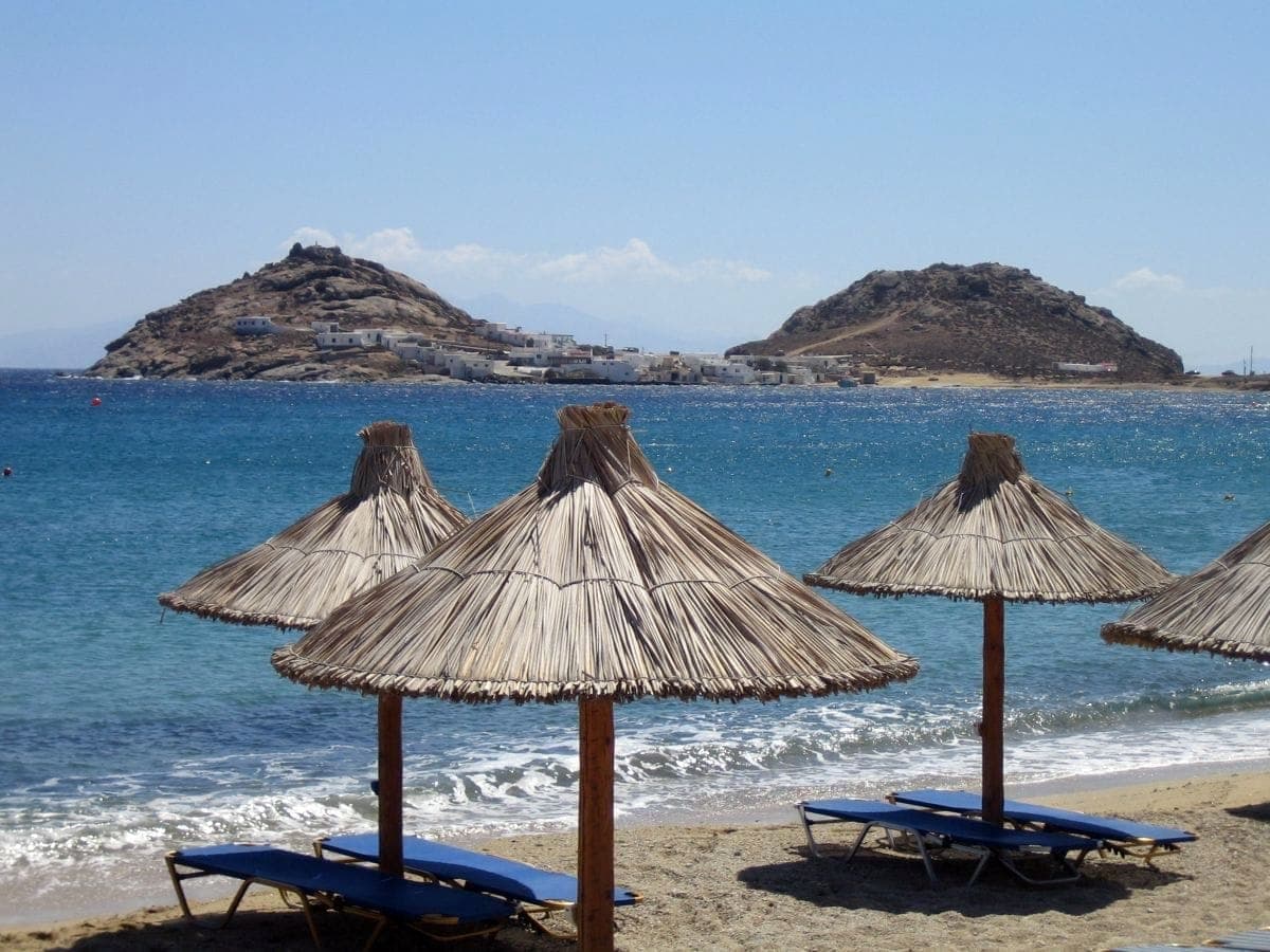Strohgedeckte Strandschirme und blaue Liegestühle am Elia Beach, Mykonos, mit klarem türkisfarbenem Wasser und felsigen Hügeln im Hintergrund.