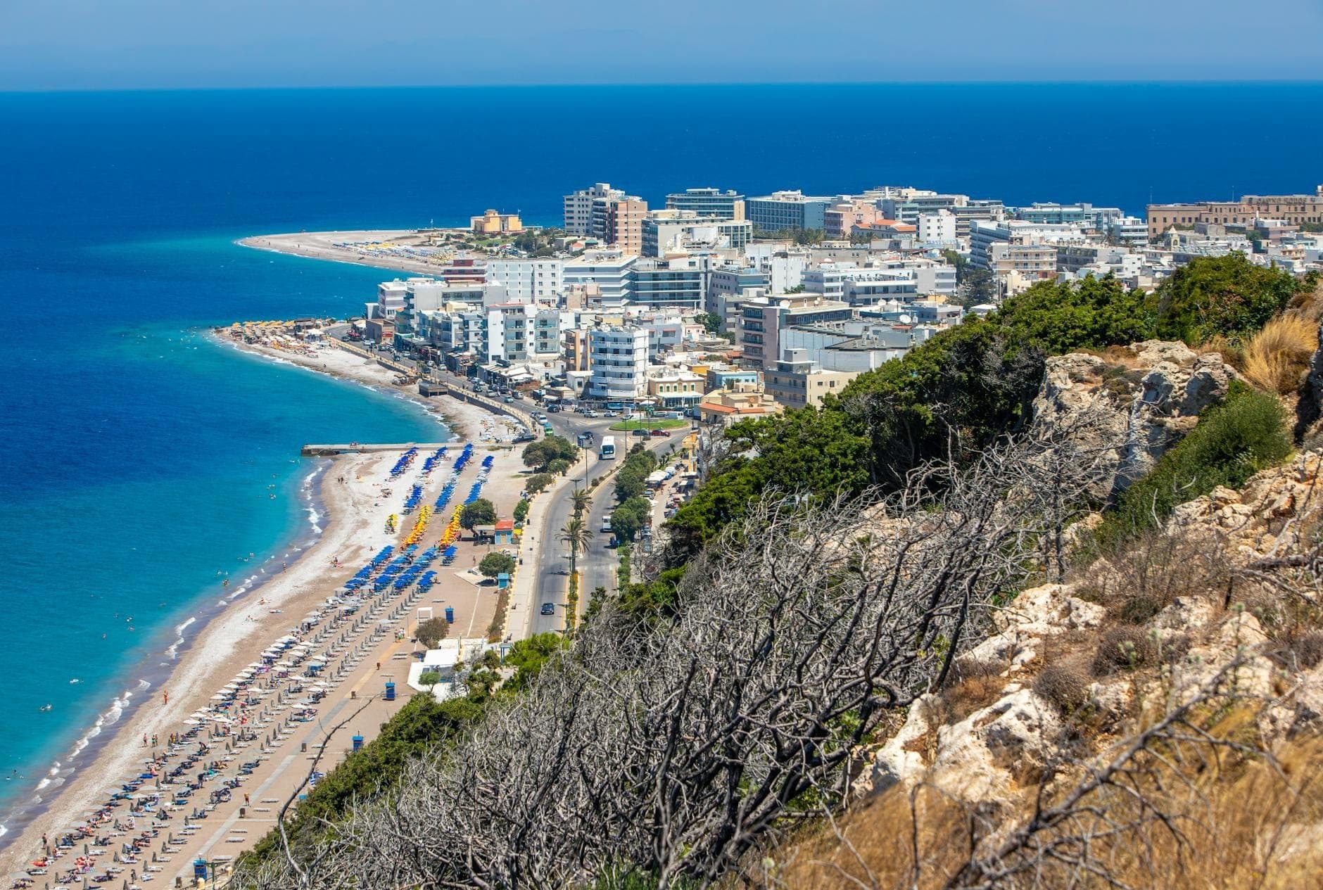 Vue aérienne panoramique de la plage d'Elli à Rhodes, avec ses eaux turquoise, ses parasols colorés, une route en courbe et les bâtiments de la ville qui rejoignent le rivage.