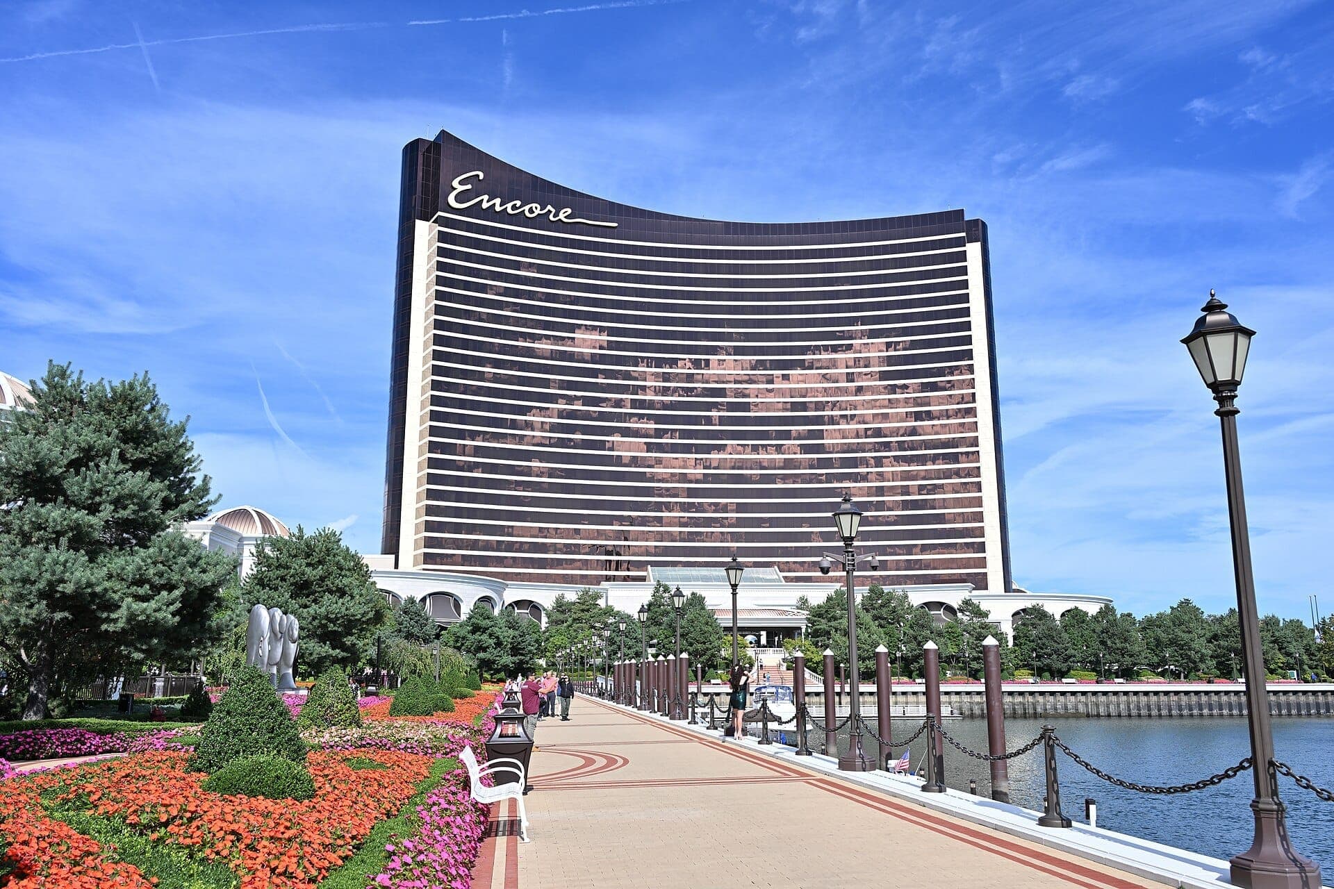 L'hôtel de luxe Encore Boston Harbor s'élève derrière des jardins colorés et une promenade le long de la Mystic River sous un ciel bleu dégagé.