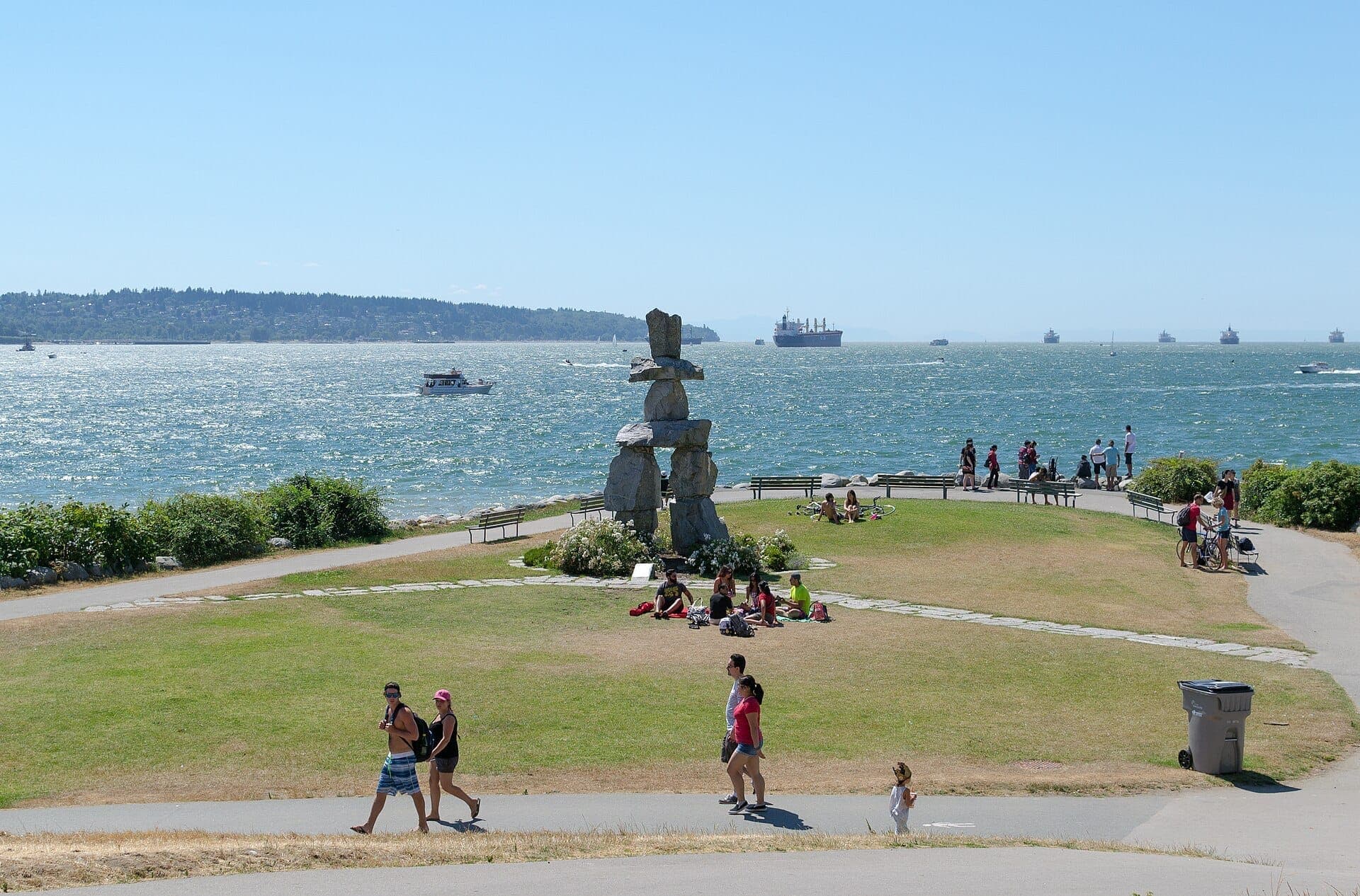 ผู้คนพักผ่อนใกล้รูปปั้น Inukshuk อันโด่งดังที่ English Bay Beach พร้อมน้ำทะเลระยิบระยับและเรือในพื้นหลังในวันที่แดดส่อง