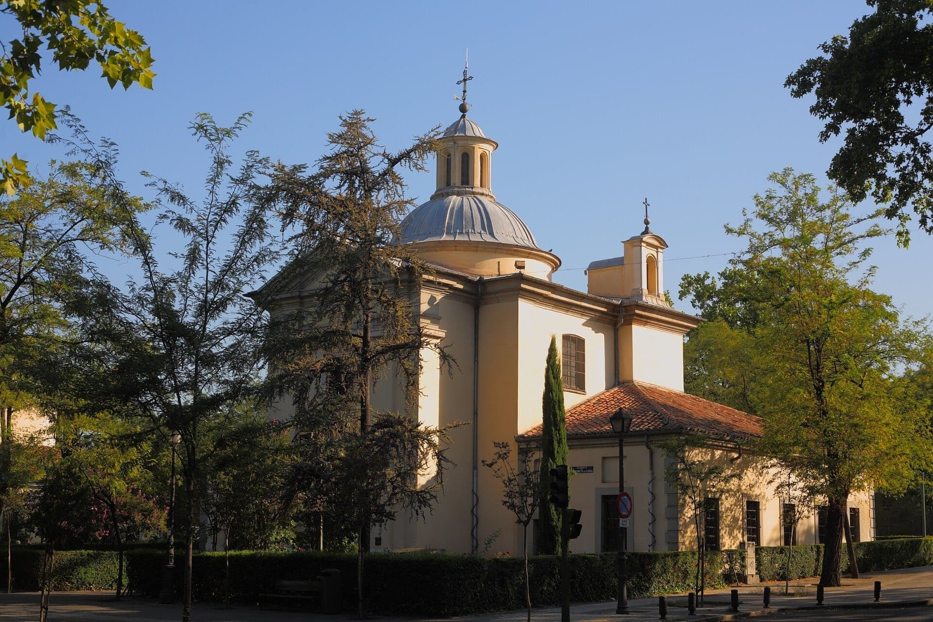 วิวของ Ermita de San Antonio de la Florida พร้อมโดมแบบนีโอคลาสสิก ด้านหน้าสีเหลืองอ่อน และต้นไม้สีเขียวโดยรอบในวันที่ท้องฟ้าแจ่มใส