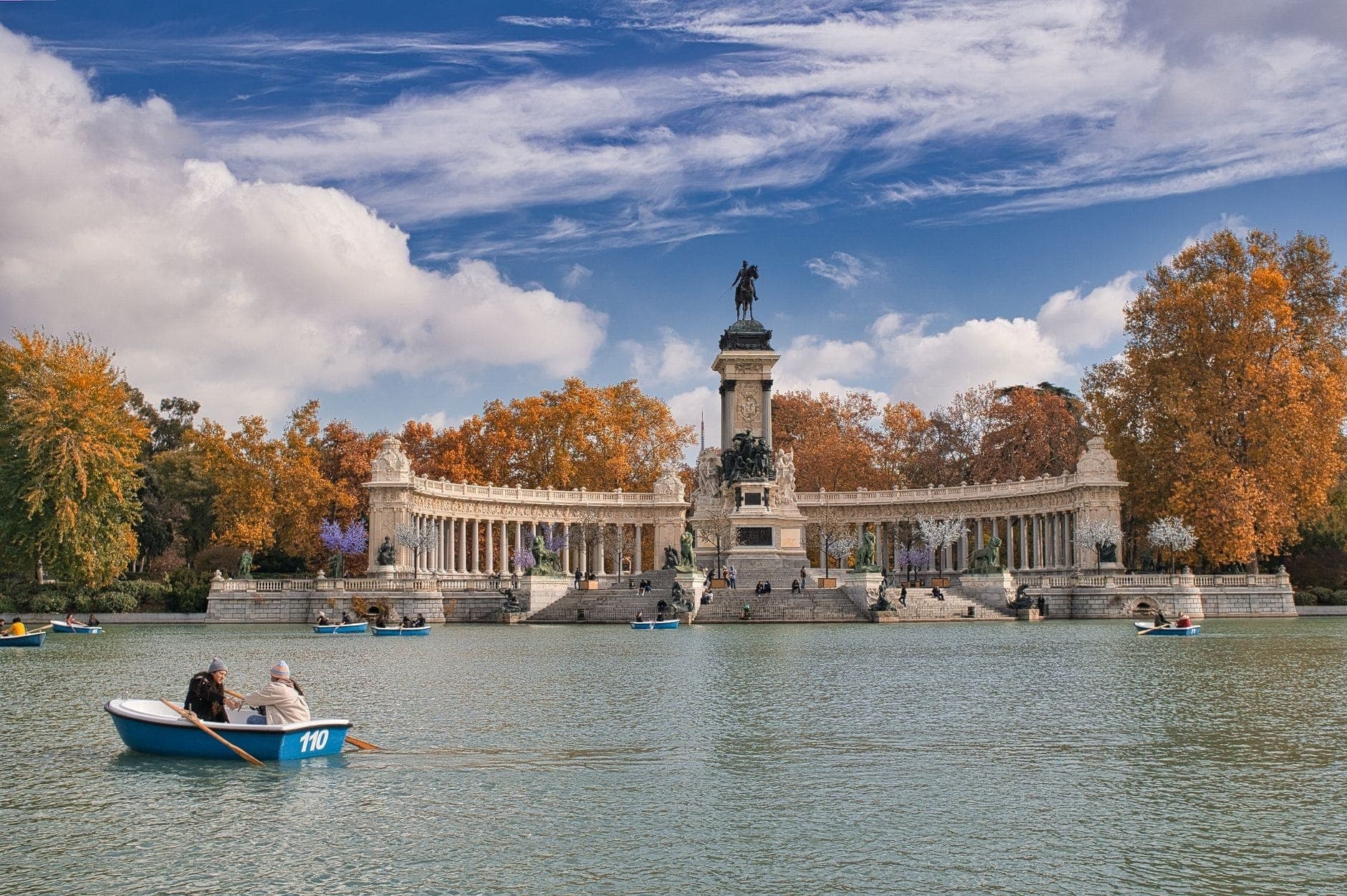 วิวทะเลสาบ Estanque Grande del Retiro กับคนพายเรือ ผสมกับฉากอนุสรณ์สถาน Alfonso XII ท่ามกลางต้นไม้เปลี่ยนสีฤดูใบไม้ร่วงและท้องฟ้าเมฆตระการตา