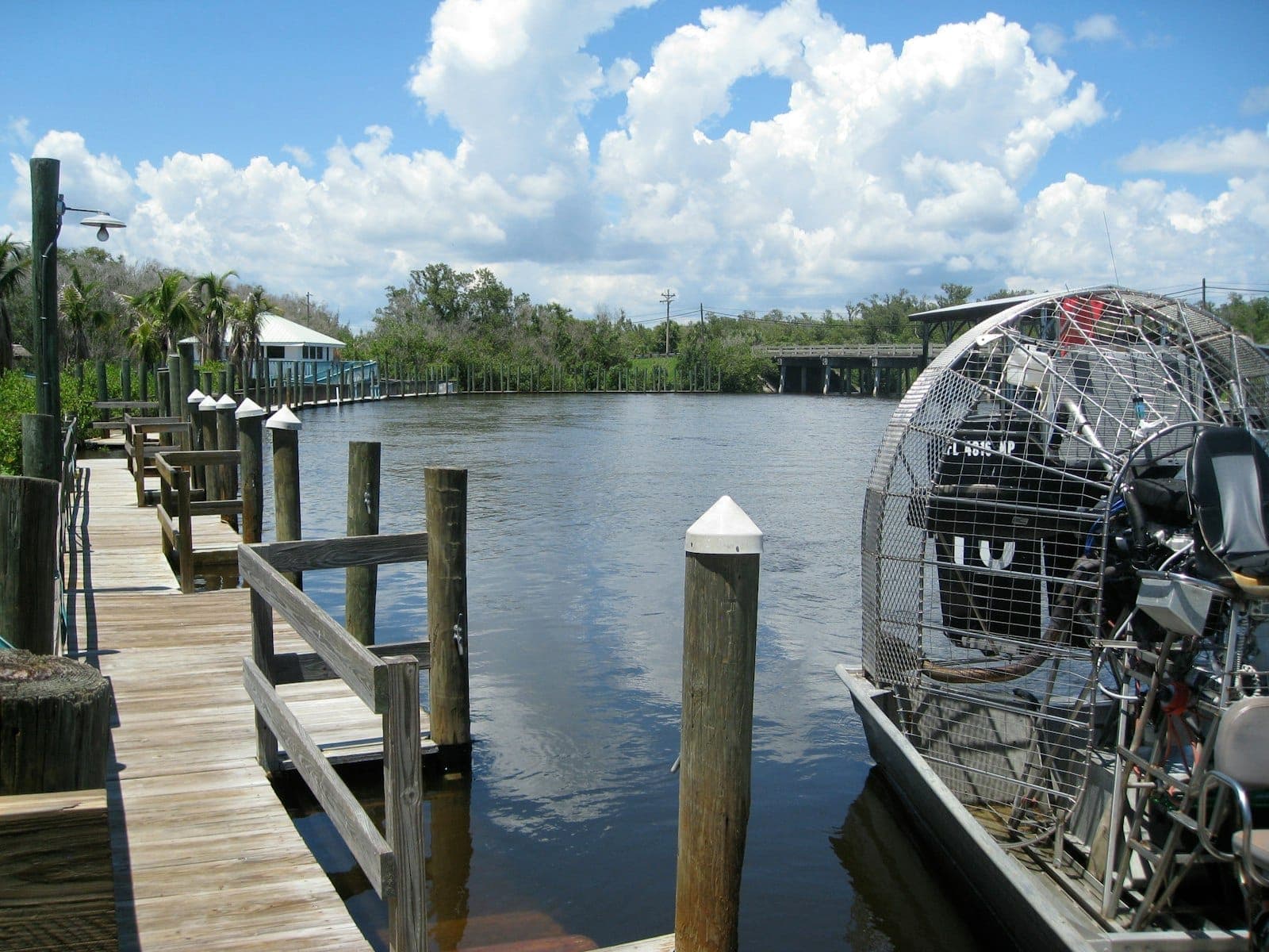 A classic Everglades airboat docked beside a wooden pier under blue skies, with lush greenery and calm water reflecting clouds.