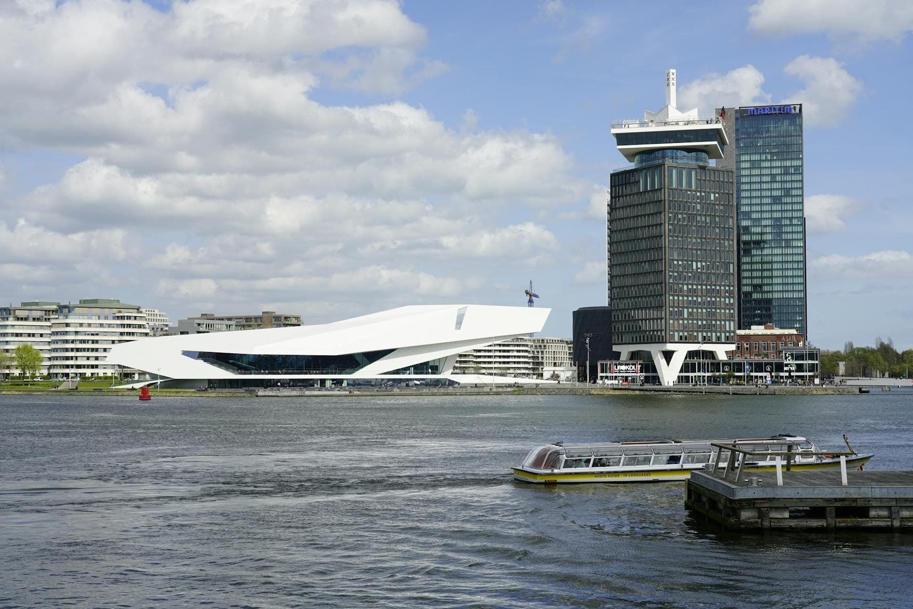 Das markante weiß-kantige Gebäude des Eye Filmmuseums und der A'DAM Tower, von der anderen Seite des IJ unter einem teils bewölkten Himmel in Amsterdam fotografiert.