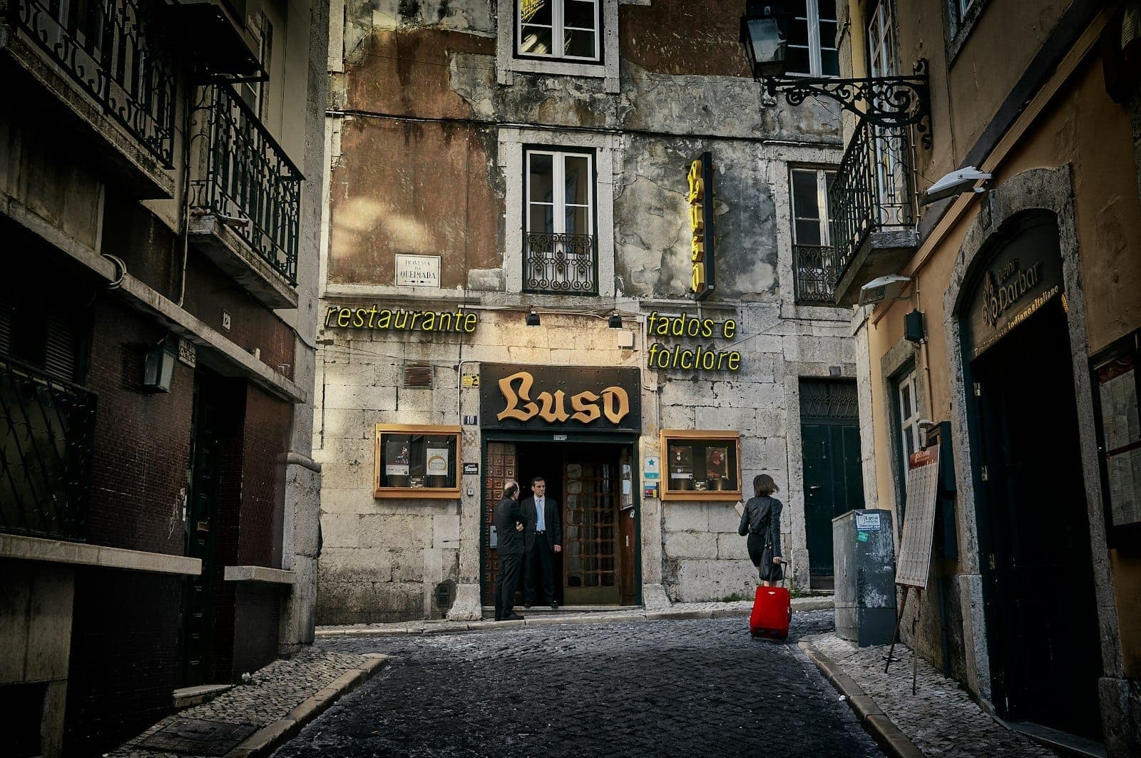 Rua estreita de Lisboa à noite em frente à entrada do 'Luso', um restaurante de fado, com pessoas e letreiro iluminado.
