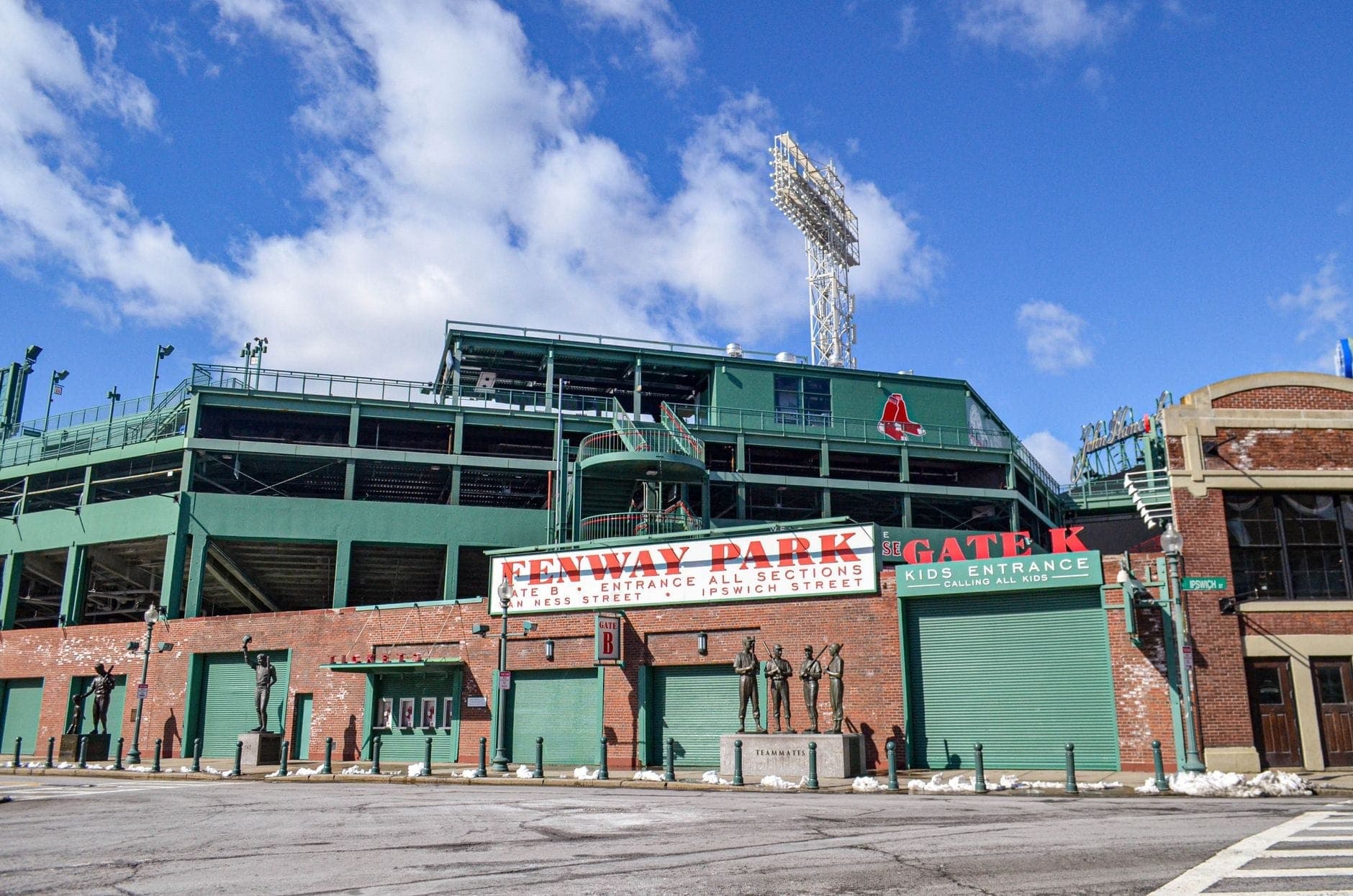 Façade extérieure de Fenway Park avec ses briques rouges caractéristiques, ses grilles vertes, l'enseigne Fenway Park et un ciel bleu dégagé.