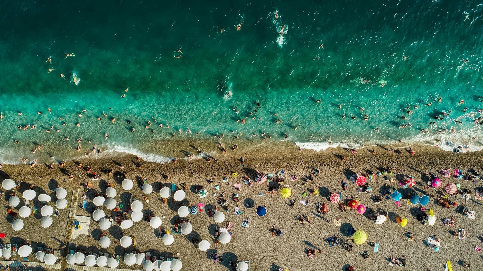 Aerial view of a crowded Antalya beach with sun umbrellas and swimmers, showing vibrant summer activity.
