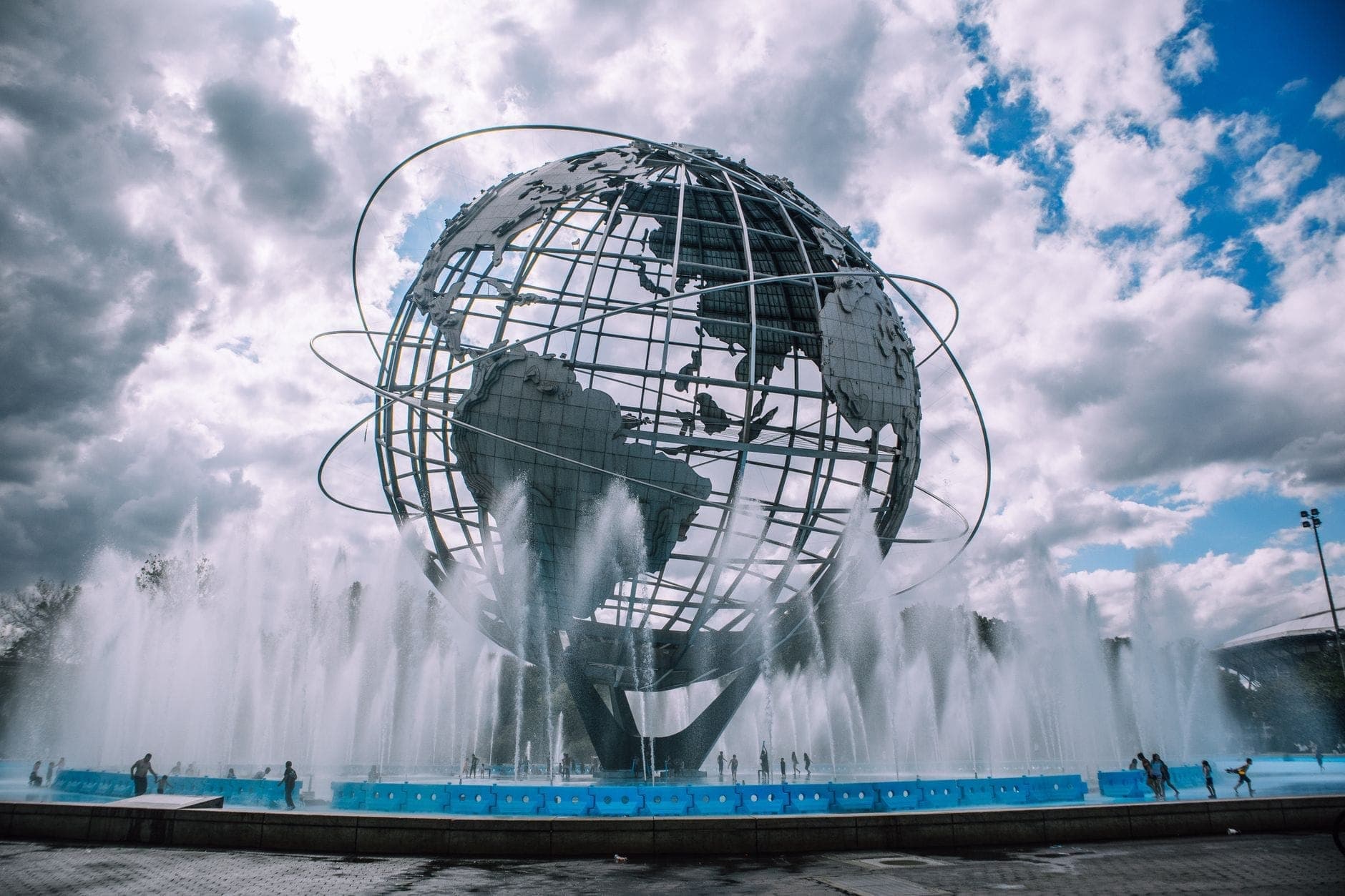 Unisphere โลกใบใหญ่ล้อมด้วยน้ำพุใน Flushing Meadows Corona Park ใต้ท้องฟ้ามีเมฆและผู้คนเดินเล่นโดยรอบ