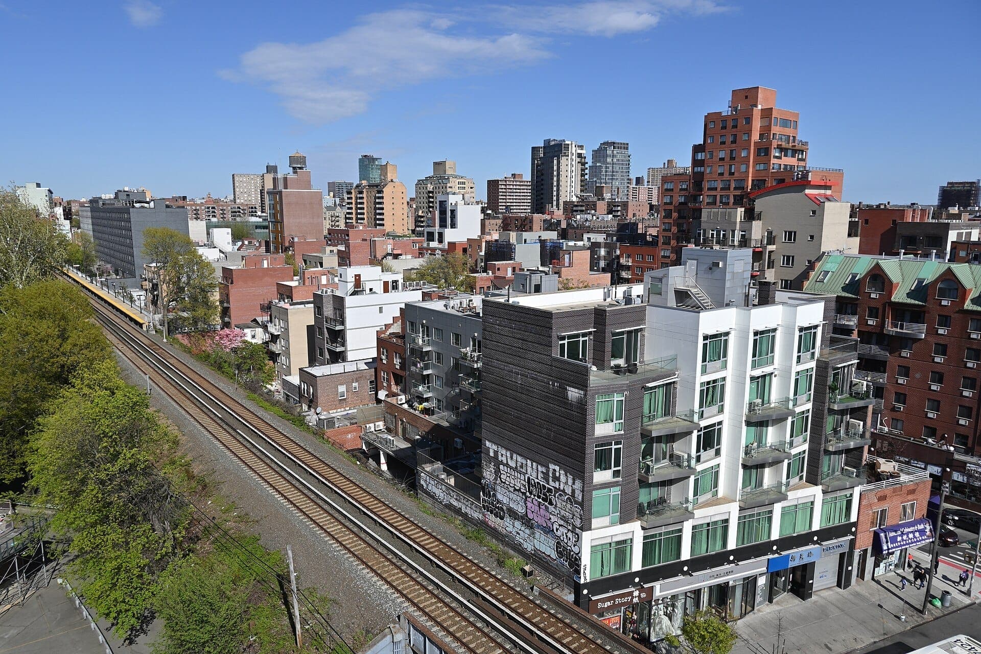 วิวกลางวันที่สว่างสดใสของ Flushing, Queens แสดงให้เห็นอาคารอพาร์ตเมนต์สมัยใหม่ ต้นไม้ร่มรื่น และรางรถไฟใต้ท้องฟ้าสีฟ้า