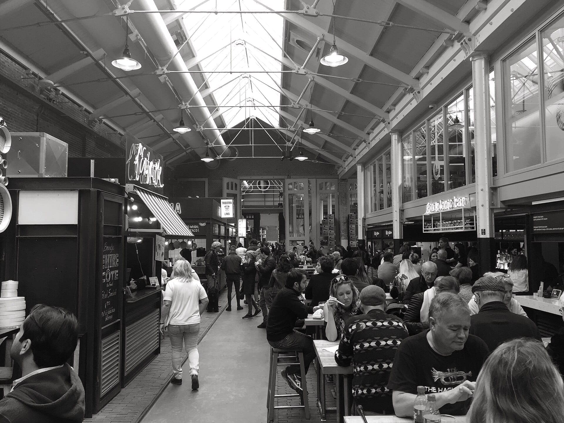 Innenansicht der Foodhallen Amsterdam mit hohen Decken, Industriearchitektur, Essensständen und Besuchern an Gemeinschaftstischen in lebhafter Atmosphäre.