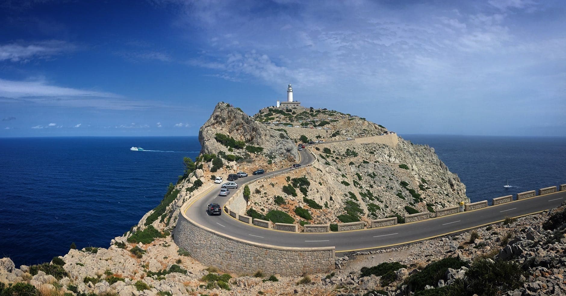 Eine kurvenreiche Küstenstraße führt zum historischen Leuchtturm Formentor auf schroffen Klippen, mit am Straßenrand geparkten Autos entlang der malerischen Mittelmeerküste auf Mallorca.
