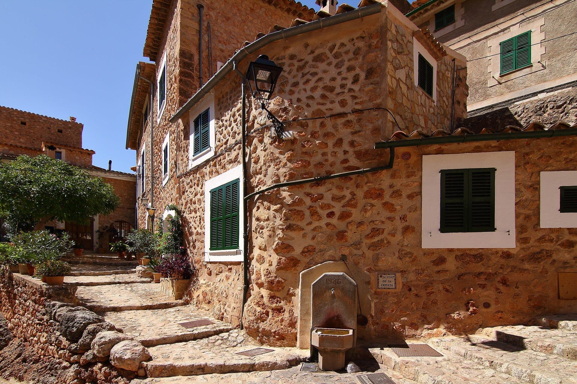 Steinhäuser mit grünen Fensterläden säumen eine sonnige Kopfsteinpflasterstraße in Fornalutx, Mallorca, mit Topfpflanzen und einem alten Brunnen.