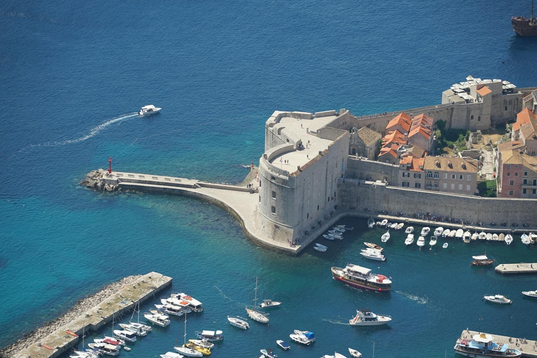 Luftaufnahme des Forts des Heiligen Johannes am Eingang von Dubrovniks Altem Hafen, umgeben von klarem blauem Wasser und kleinen Booten, mit Stadtmauern und Dächern im Hintergrund.