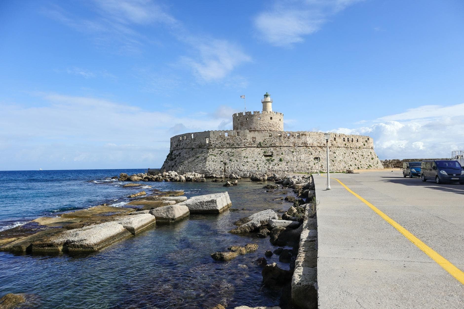 Le fort Saint-Nicolas à l'extrémité de la jetée du port de Mandraki, cerné par une mer et un ciel bleus, avec une vue dégagée sur son bastion rond et son phare.