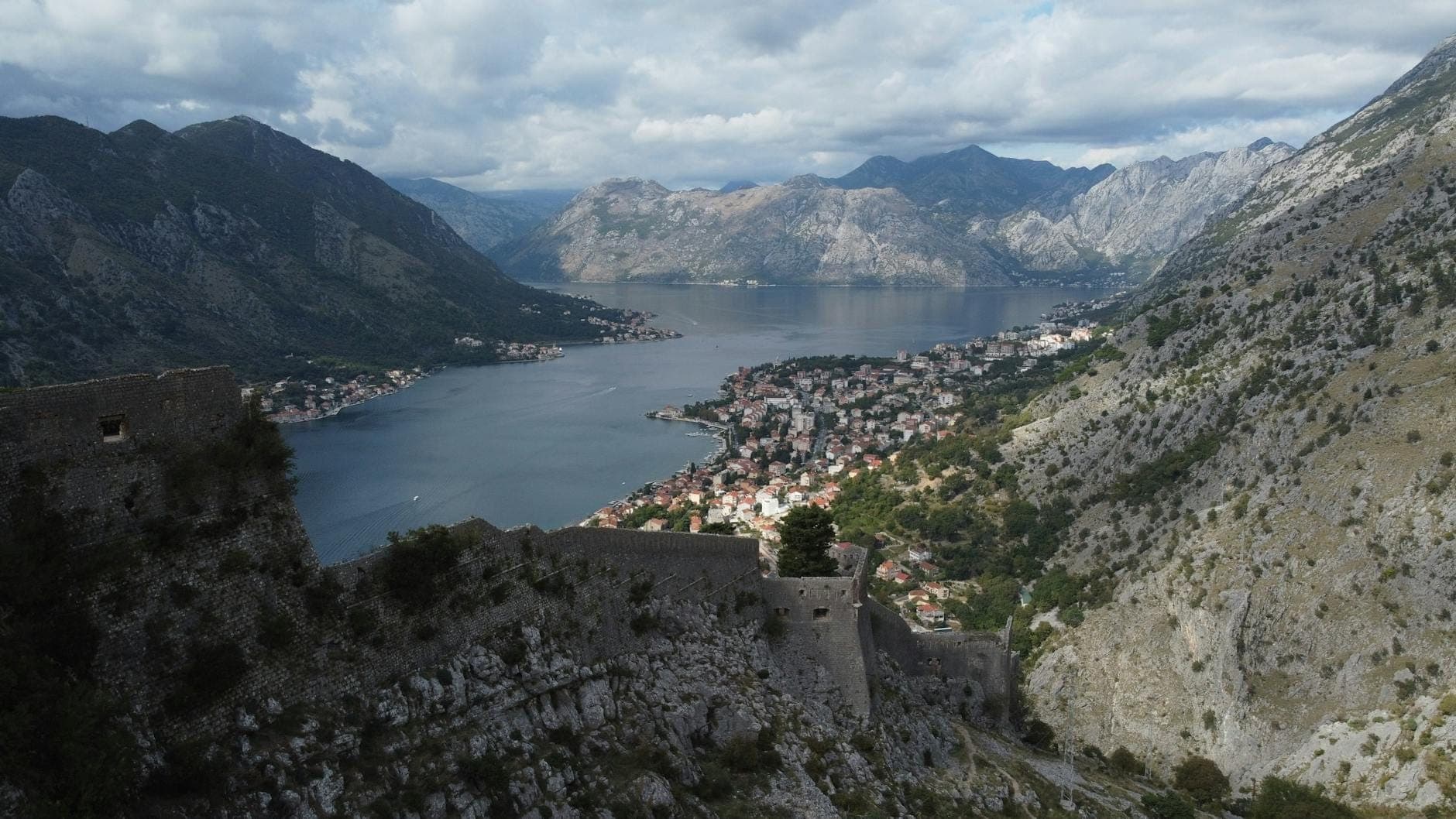 Vue panoramique depuis la forteresse de San Giovanni sur la vieille ville de Kotor, la baie et les montagnes environnantes sous un ciel partiellement nuageux.